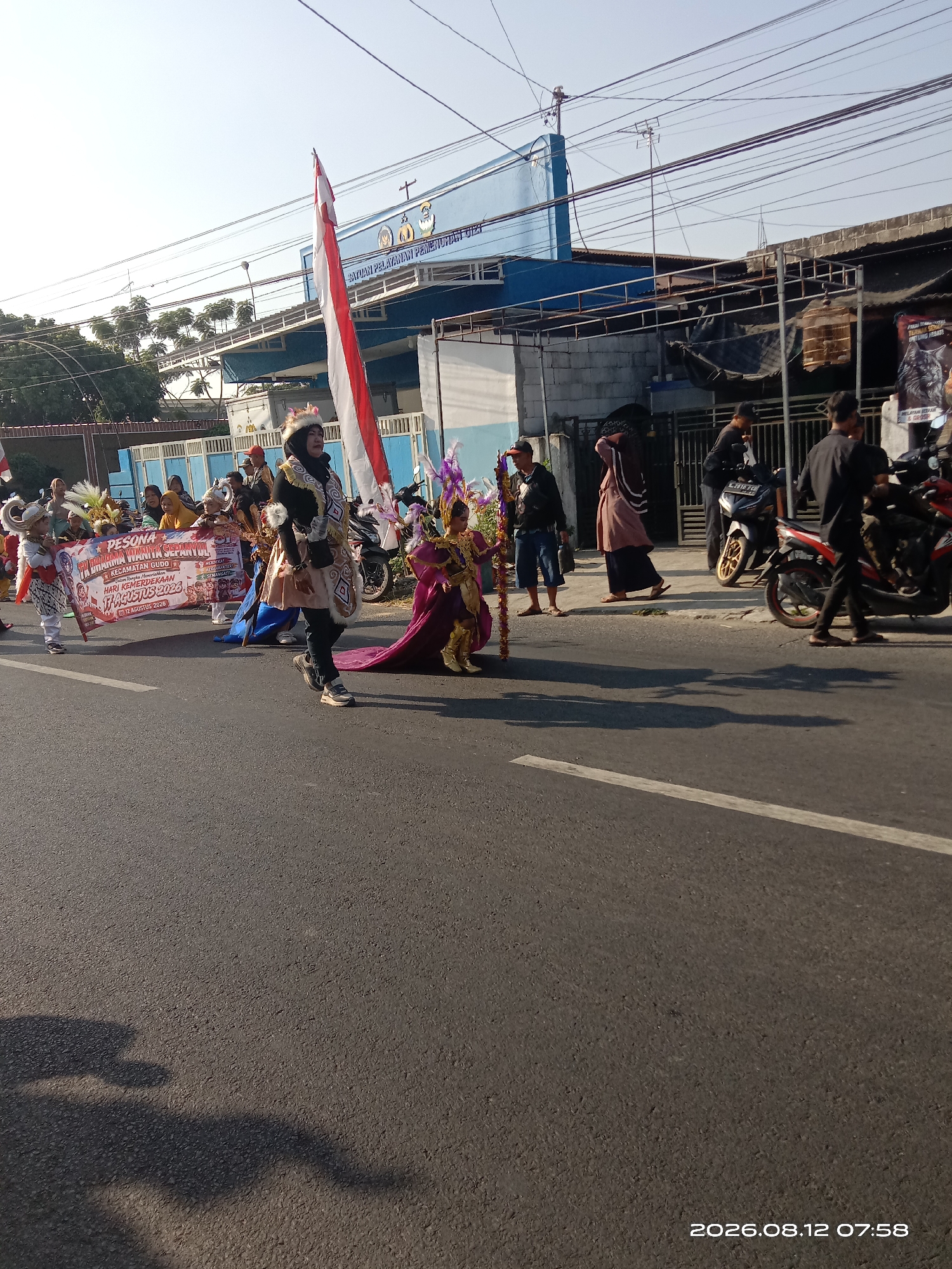 The image captures a street scene in Gudo, Indonesia, on August 12, 2026, at 07:58. The foreground shows a paved road with a white line, and shadows cast from the right. A parade is in progress.
In the center of the image, a group of people are walking, some in traditional or festive attire. A prominent figure in the foreground on the right wears a black outfit with a furry trim and a light-colored skirt, holding a staff with colorful ornaments. To their left, a child in a purple and gold costume with a long train is walking.
Further left, a group of individuals are holding a banner that reads "PESONA KARNA WANITA SEPANYOL KECAMATAN GUDO HARI KEMERDEKAAN 17 AGUSTUS 2023." The banner is adorned with illustrations and text. Several other people in the parade are wearing decorative headwear and colorful clothing. A Indonesian flag is being carried, with red and white stripes.
The background features a light blue building with signage that reads "SATUAN PELAYANAN PEMERINTAH". There are also several motorcycles parked on the side of the road, and a few people standing and observing the parade. Trees and overhead electrical wires are also visible. The overall scene suggests a community event or celebration.