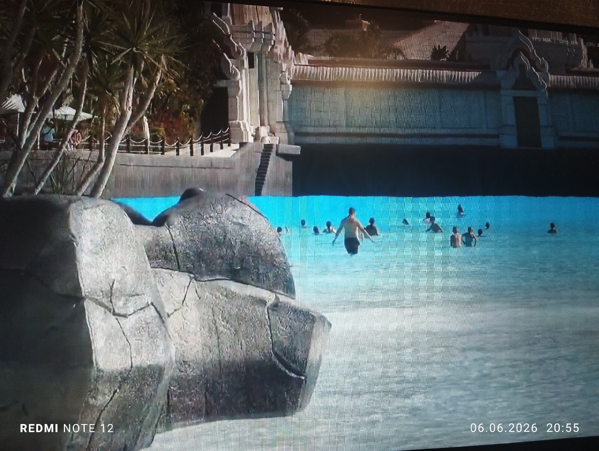 This image is a photograph captured by a "REDMI NOTE 12" smartphone on June 6, 2026, at 20:55, displaying a digital screen. The screen exhibits performance metrics "FPS N/A GPU 0% CPU 2%" in the upper right quadrant, indicating the original content is a rendered scene, likely from a video game or simulation.
The scene depicted is a large, elaborately themed water park within the city of Burgas, Bulgaria, presented at dusk or night. In the foreground, large, grey, artificial rock formations with visible cracks and rough textures occupy the left and center-left, partially framing a body of water. The water in the immediate foreground and midground appears light blue, exhibiting subtle surface ripples.
The midground features a spacious wave pool or large swimming pool. Approximately 10 to 15 individuals are present in the water. One male subject is centrally positioned, visible from the waist up, wearing dark swim trunks, and appears to be walking through the water. Other subjects are further back, partially submerged, their specific actions indistinct due to distance and image resolution.
The background is dominated by an extensive, multi-tiered architectural complex designed in a style reminiscent of ancient temple ruins, possibly Southeast Asian or Mesoamerican in influence. Key architectural details include vertical pilasters, ornate cornices, and textured wall sections. A dark, arched entrance is visible on the lower left of the structure. A staircase descends from a higher platform to a lower one on the left. Decorative railings with spherical finials line various ledges. A horizontal channel of dark water runs across the mid-section of the background structure, forming a waterfall over a textured wall below. To the far left, a cluster of tall, slender-trunked plants, likely palm trees, with dark fronds, partially obscure the architecture. White parasols are visible beneath these trees, indicating a relaxation area. The sky above the structures is dark, consistent with the recorded time.