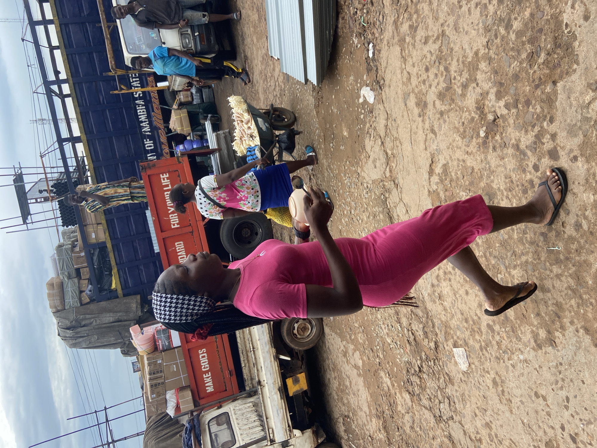 This media file depicts a scene in Ogbunike, Nigeria. A young woman in a bright pink dress and black flip-flops is the central subject, her gaze directed upwards and to the left. She has braided hair adorned with a black and white patterned headscarf. Behind her, a blue truck filled with goods is parked. The truck has text on its side that reads "ANY OF ANAMBRA STATE." To the left of the blue truck, a red truck is visible, also carrying cargo. Text on the red truck's side states "MAKE GOODS." In the background, other individuals are present, some appearing to be involved in activities related to the trucks and market. One person, wearing a blue shirt, stands near the blue truck, while another, in a dark top, is further in the background. A wheelbarrow loaded with what appears to be produce is also visible in the mid-ground. The ground is rough and appears to be dirt or gravel. The sky is cloudy, suggesting a potentially overcast day. The overall atmosphere suggests a busy marketplace or loading area.