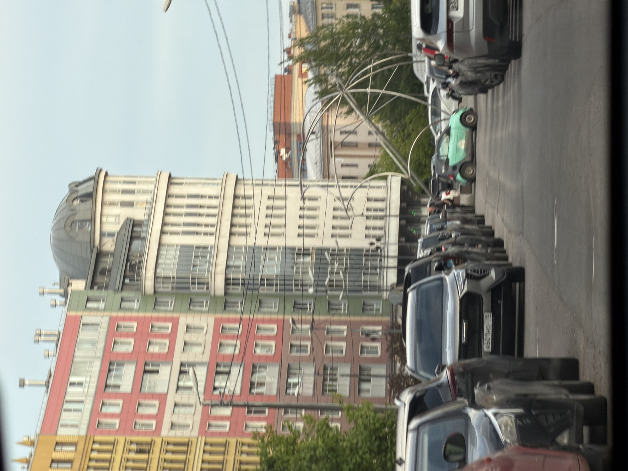 The image displays a vibrant urban street scene in Saint Petersburg, Russia, characterized by diverse architectural styles and numerous parked vehicles under a bright, possibly overcast sky.
In the background, several multi-story buildings dominate the cityscape. A prominent cream-colored building features a distinctive dark grey dome on its roof and numerous evenly spaced windows, suggesting a classic or grand architectural style. Adjacent to it, a colorful building stands out with its facade divided into horizontal sections of red, green, and yellow, likely indicating a more modern residential or commercial structure. Further back, hints of other buildings with terracotta or light-colored facades are visible.
Along what appears to be the right side of a city street, a continuous line of cars is parked, extending into the distance. A light green Smart car is clearly visible among them. Closer to the foreground, a modern silver SUV, identified by its grille, bears a Russian license plate with the registration `О501МР 98`. The region code `98` on the license plate specifically denotes Saint Petersburg, confirming the provided location context. Other vehicles, including an older dark-colored car and a red car, are also partially visible in the foreground and stretching down the street.
Numerous power lines or cables crisscross the upper portion of the scene, adding a layer of urban complexity. Above the parked cars, a large, ornate metal archway or sculptural element spans across the street, possibly serving as a decorative installation or part of the city's public art. Scattered trees and green bushes indicate some natural elements integrated into the urban landscape. The overall impression is that of a bustling city environment during daytime. No people are clearly visible in the frame.