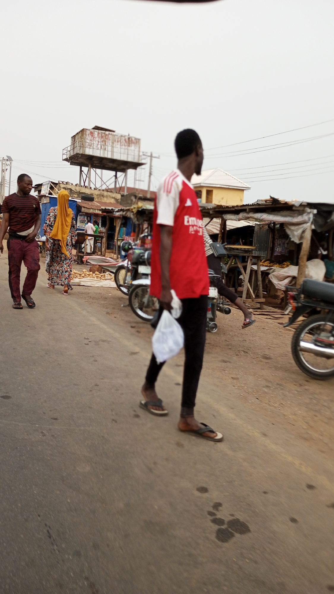 The scene is a street in Adankolo, Nigeria. A man in a red and white shirt, black pants, and sandals is walking away from the camera, holding a white plastic bag. Two other people are walking in the opposite direction: a man in a maroon striped shirt and maroon pants, and a woman in a floral dress and yellow headscarf. Parked motorcycles line the side of the street near informal market stalls with corrugated metal roofs. An elevated metal water tank is visible in the background. The road is paved and has patches of dark stains.
