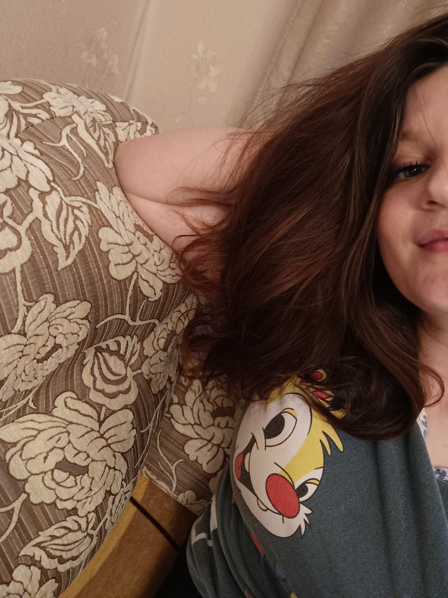 A woman is resting on a couch, supporting her head with her arm. She has brown hair, dark eyelashes, and part of her face is visible. She is wearing a blue t-shirt with a cartoon image on it. The couch has a floral pattern. There is no information available to determine the location of the image in the city of Tobolsk, Russia.