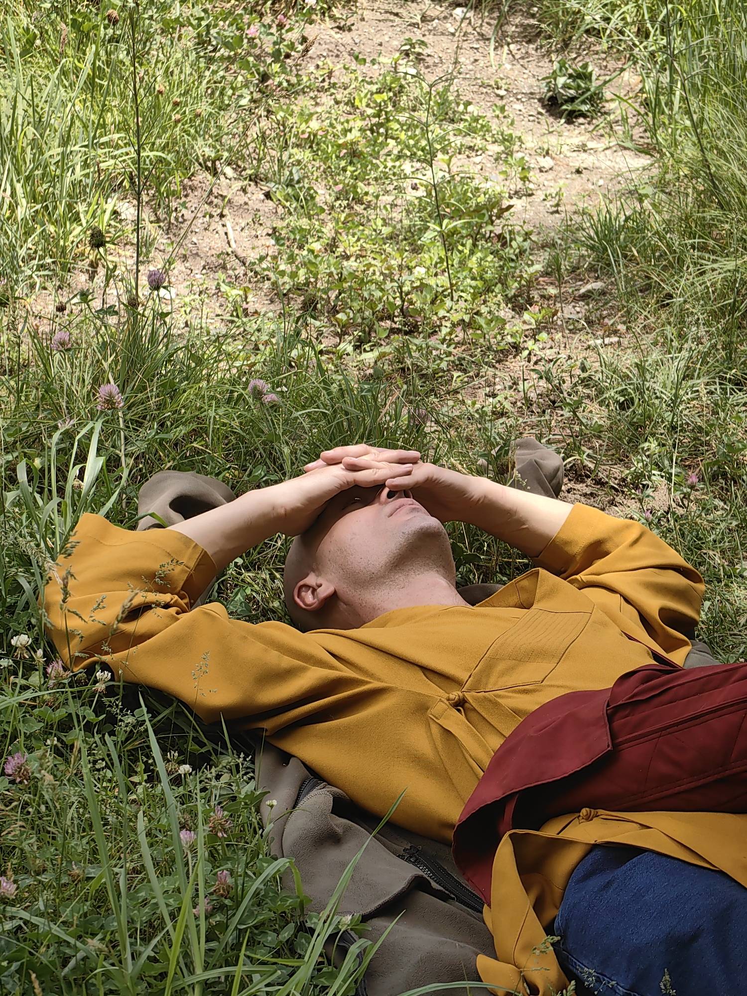 A person lies in a grassy field with their hands over their eyes. They are wearing a yellow tunic and blue jeans. The field is covered in green grass and small pink and white flowers. The person's head is bald, and they appear to be resting or sleeping. The scene is set in Barheta, India, specifically in a natural environment. The lighting suggests it is daytime, possibly a sunny day given the shadows. The overall impression is one of peaceful repose in a natural setting.