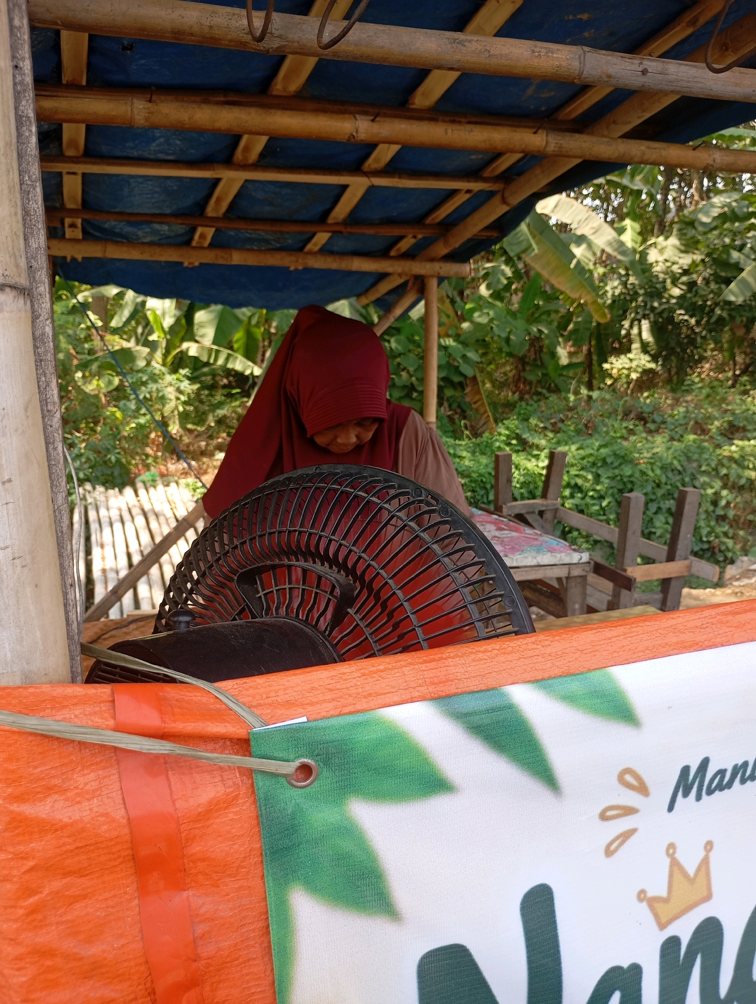 A woman wearing a maroon hijab and a brown shirt is seated under a bamboo and blue tarpaulin roof, tending to a black oscillating fan. Behind her, a lush green background suggests an outdoor environment with trees and foliage. In the foreground, an orange tarp with green leafy graphics and text is visible. The scene appears to be in Babelan, Indonesia, possibly at a market or roadside stall.