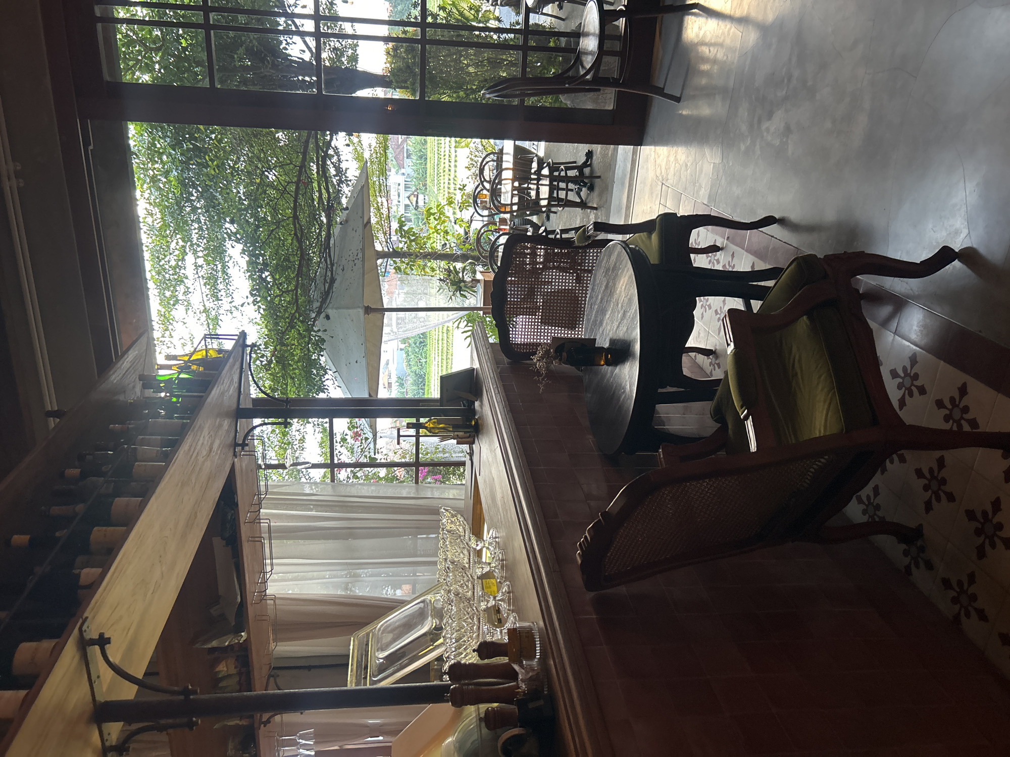 This image depicts the interior of a cafe or restaurant with an outdoor seating area, likely located in North Kuta, Indonesia, given the lush green rice fields visible through the open doorways.
Inside the establishment, a wooden bar is prominent, stocked with bottles and glasses on shelves above. Crystal bowls are displayed on the bar counter. In the foreground, two ornate chairs with green upholstery flank a dark wooden table. The floor inside is tiled with a patterned design.
The outdoor area features more tables and chairs, some under a large white umbrella. Beyond the seating, a verdant landscape unfolds, characterized by flooded rice paddies and scattered trees, bathed in natural light suggesting either early morning or late afternoon. The architecture suggests a fusion of traditional and modern elements, with wooden beams, large glass doors, and an open-air feel. No people are visible in the image.