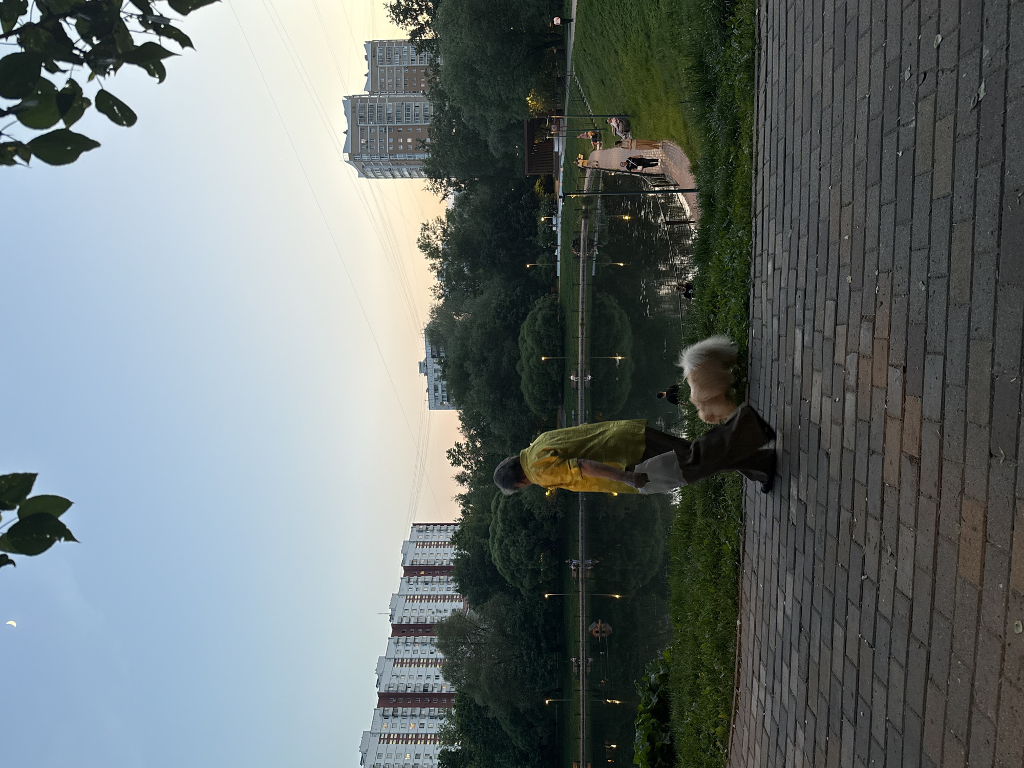 The image shows a park setting, likely in Moscow, Russia, based on the architecture of the residential buildings in the background. A brick walkway occupies the foreground on the right side of the image. A person in a light-colored jacket stands next to a dog with fluffy, light-colored fur. They are positioned near a pond or lake, which reflects the surrounding trees, buildings, and streetlights. Other individuals can be seen in the distance near the water's edge. The sky appears to be at dusk or dawn. Vegetation includes trees and grass around the pond. The buildings in the background are tall, multi-story apartment complexes.