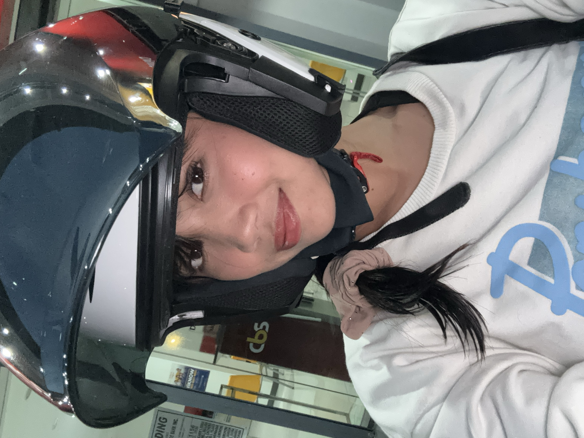 A young woman wearing a motorcycle helmet with a dark visor smiles at the camera. She is wearing a white long-sleeved shirt with a light blue logo on the front. The background appears to be inside a building with fluorescent lighting. Signs are visible in the background, one with the text "ENDING" and another with "cbs." The overall impression is a selfie taken before or after a motorcycle ride.
