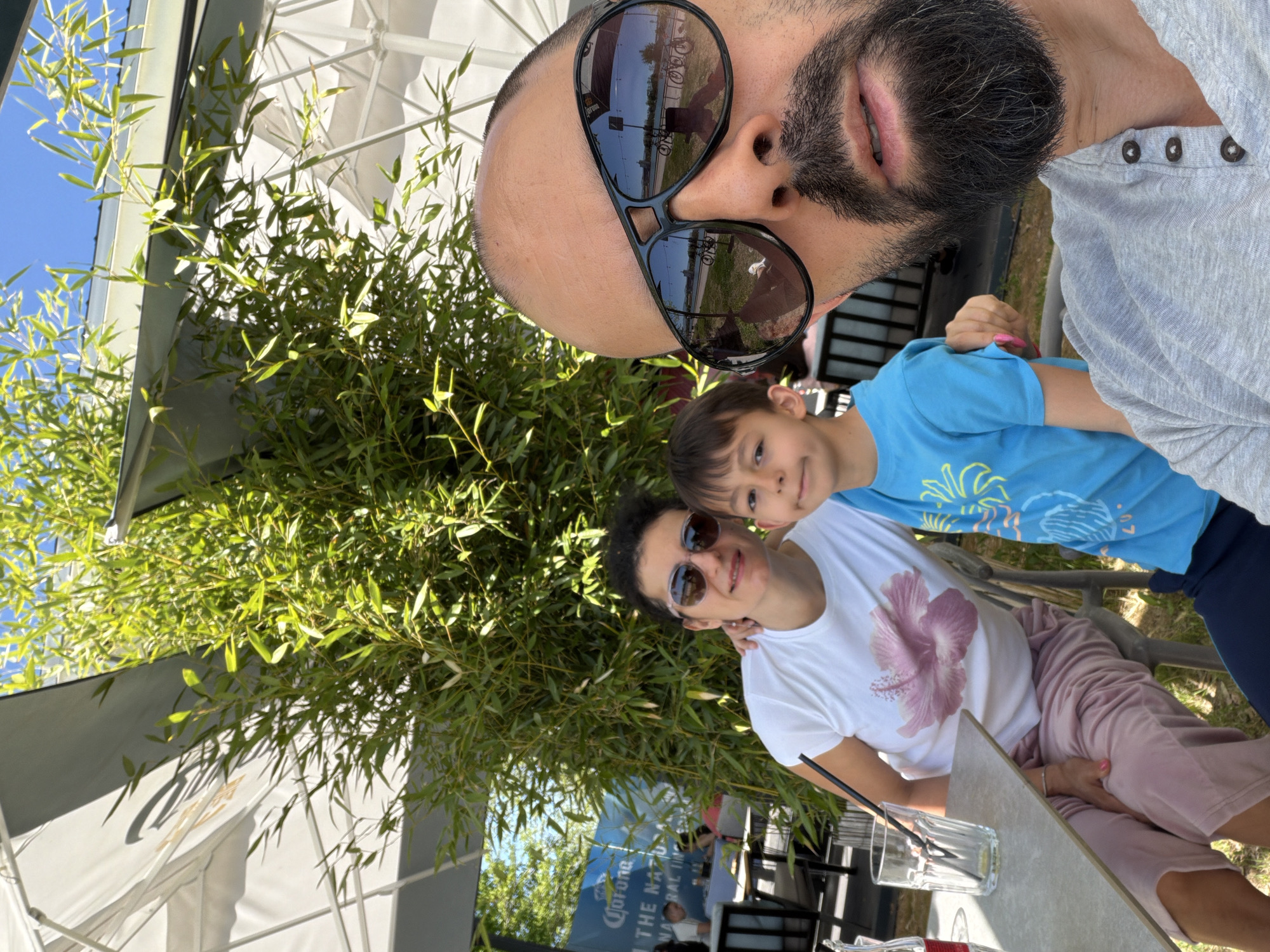The image shows a selfie of a man, a woman, and a child, seemingly taken at an outdoor cafe in Plovdiv, Bulgaria. The man at the top of the frame has sunglasses reflecting a street scene, a beard, and is wearing a grey button-down shirt. The woman and child are positioned lower in the frame; the woman is wearing sunglasses and a white t-shirt with a pink flower design, and the child is in a blue shirt. A glass of liquid with straws sits on the table in front of them. In the background, there are other cafe patrons and the surrounding foliage, including bamboo. A partial Corona beer advertisement is visible in the background. Umbrellas are providing shade.