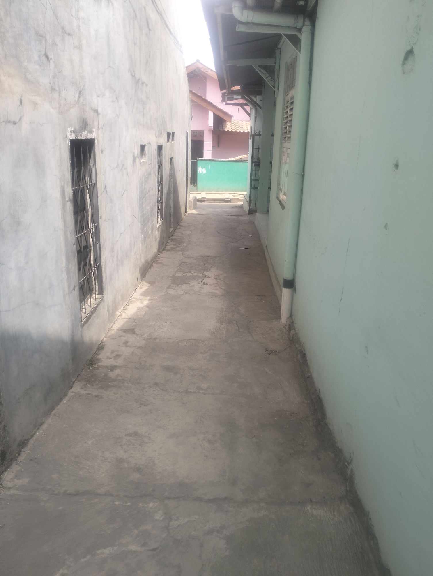 The image depicts a narrow alleyway between two buildings in Karangsoka, Indonesia. The left building has a rough, gray concrete wall with several small, barred windows. The right building is painted a pale, mint green and features a window with louvers. The ground of the alley is paved with concrete, showing signs of wear and tear with cracks and some grass growing along the edges. In the background, a pink building with a tiled roof is visible. A green, corrugated metal wall or barrier is also present in the distance. The lighting suggests it is daytime, with shadows cast by the buildings and roof overhangs. There are no people or animals visible in the scene.