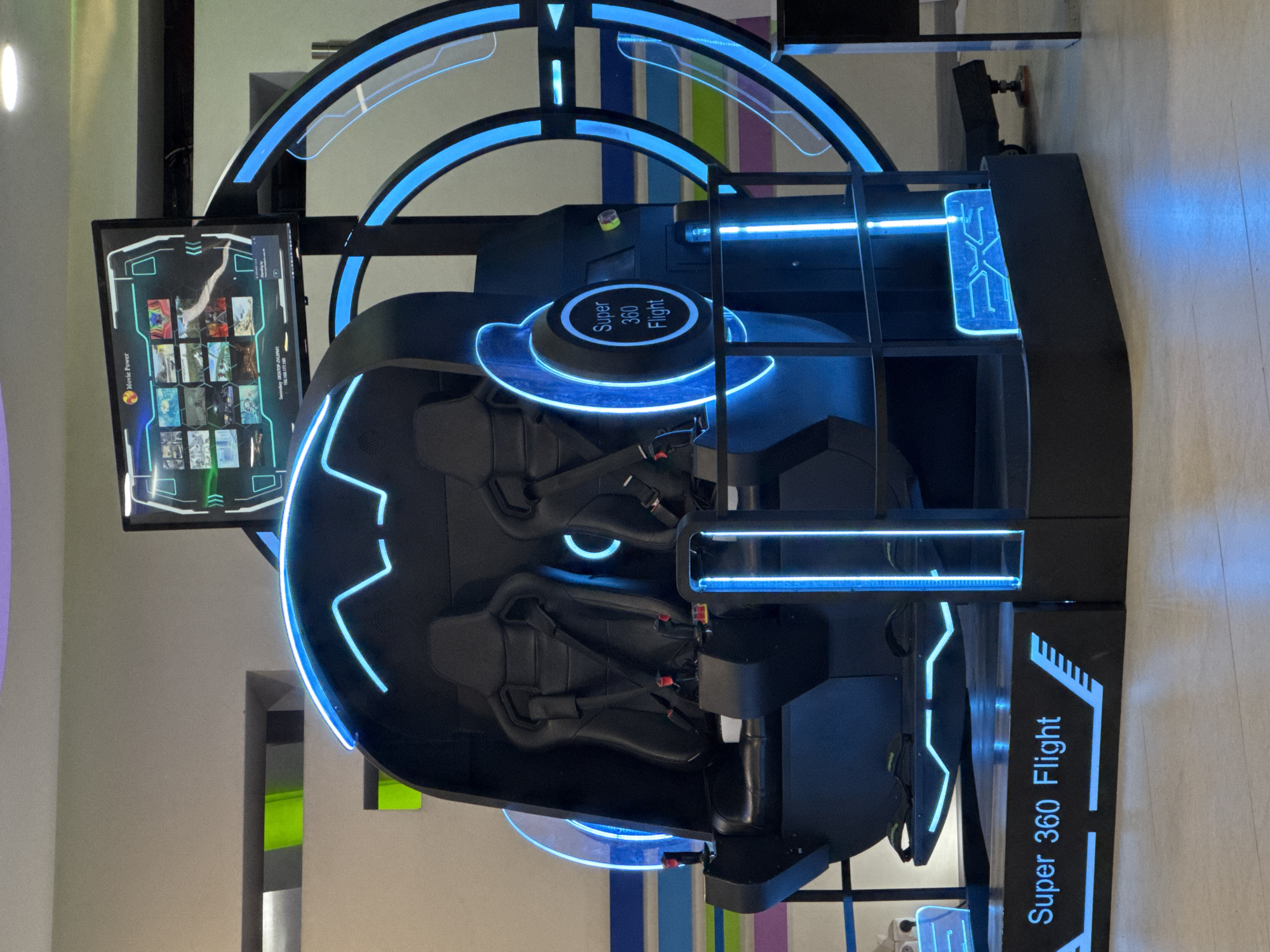 This image showcases a futuristic, dark-colored virtual reality or simulation ride, likely situated in an entertainment center or arcade in Yerevan, Armenia. The machine is predominantly black, strikingly accented by bright blue LED lighting that outlines its structural arcs, circles, and geometric patterns, imparting a modern, high-tech appearance.
At the heart of the simulator are two black bucket seats, equipped with safety harnesses, indicating a dynamic and immersive experience. A prominent central circular platform is illuminated with blue light and displays the text "Super 360 Flight." This name is reiterated on the bottom right base of the machine, accompanied by stylish arrow-like graphics.
To the left, a vertical screen functions as a user interface. It features "Movie Power" branding at the top left and presents a grid of thumbnails, likely representing various virtual experiences or movies available for selection. Partially visible text on the screen includes "Sound only," a date and timestamp ("Saturday 2023-01-28 17:01:45"), and an IP address ("IP: 192.168.1.100"). A scrollbar suggests a broader selection of content.
The simulator is currently inactive, with no people present, but appears ready for use. The setting is indoors, with light-colored walls and a light-toned floor, and hints of colorful vertical stripes in the background. The overall scene conveys an atmosphere of contemporary and engaging entertainment.