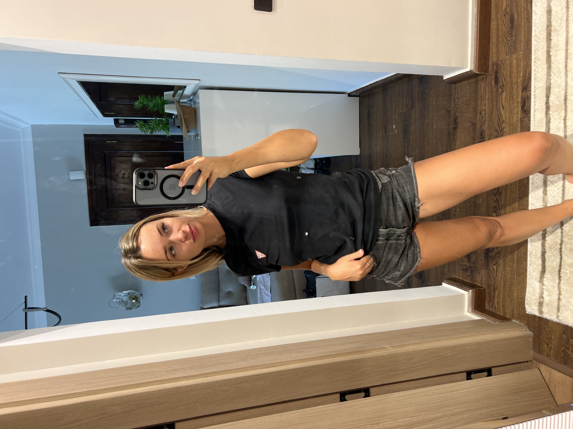 A blonde woman with light skin and a light complexion is standing in front of a mirror and taking a selfie. She is wearing a black t-shirt with a red and white triangular logo on the left chest and distressed denim shorts. The woman has light blonde hair that is cut to shoulder length. She is wearing a ring on her left ring finger. The background shows a doorway on the left and a mirror on the right. The mirror reflects a white cabinet with plants and a straw hat, as well as a dark wooden door. The flooring is dark wood. The scene appears to be an indoor residential setting.