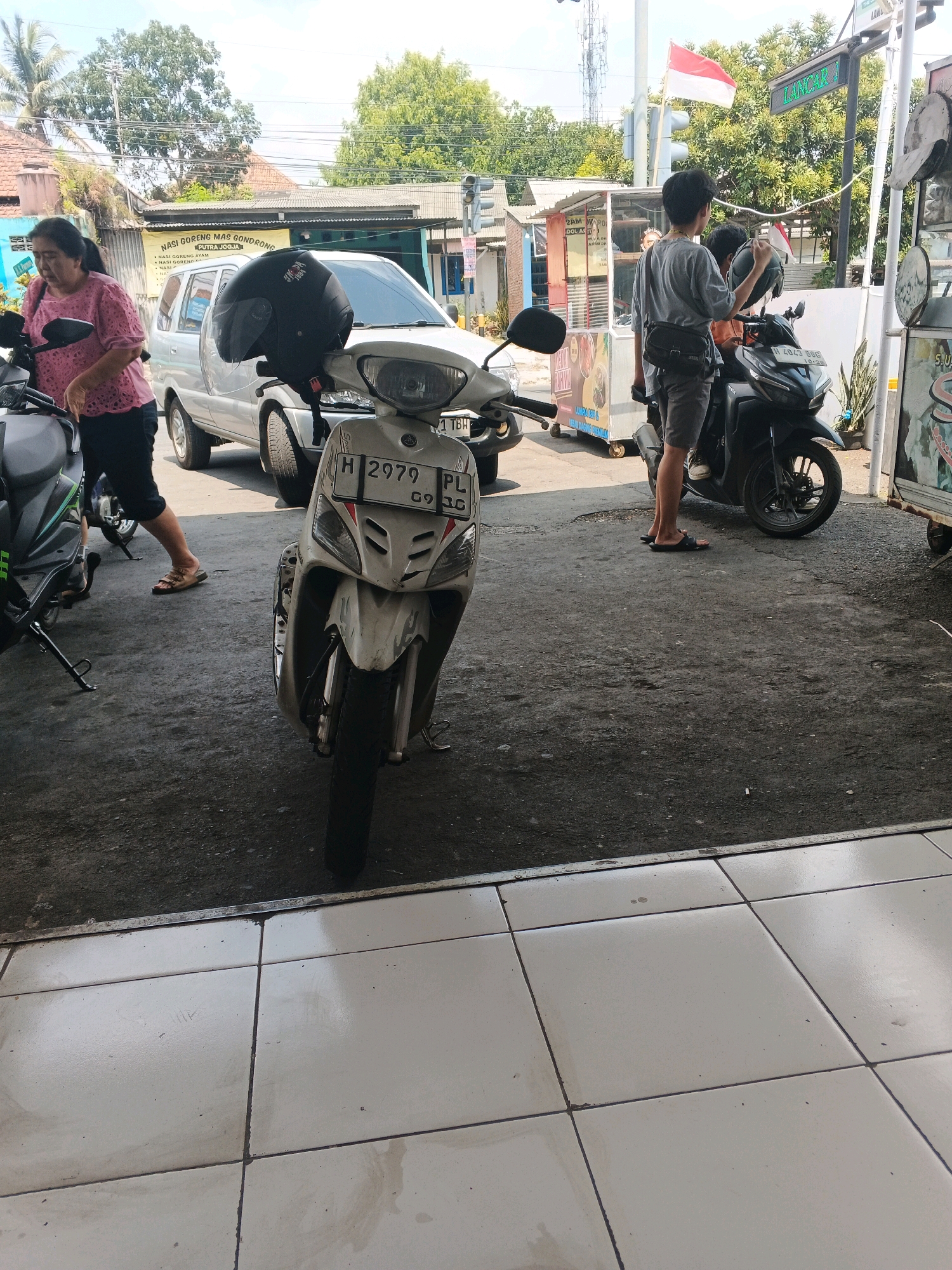 The image shows a street scene in Genuk Barat, Indonesia, with several motorcycles and a car parked on a paved surface. In the foreground, a white scooter with the license plate "H 2979 PL" is parked. To the left, a dark grey scooter is partially visible. A silver car is parked behind the white scooter. A woman wearing a pink patterned shirt and black pants is standing next to the dark grey scooter, appearing to interact with it. Further back, a young man in a grey t-shirt and dark shorts is sitting on a black motorcycle, holding a helmet. Another person is partially visible on the back of the black motorcycle. Behind the vehicles, there are buildings, including what appears to be a food stall with signage that includes the text "NASI GORENG MAS GONDRONG PUTRA JOGJA". A red and white Indonesian flag is flying from a pole. The environment appears to be an outdoor area with trees and overhead wires, suggesting a typical urban or semi-urban setting. The ground is a mixture of asphalt and tiled flooring.