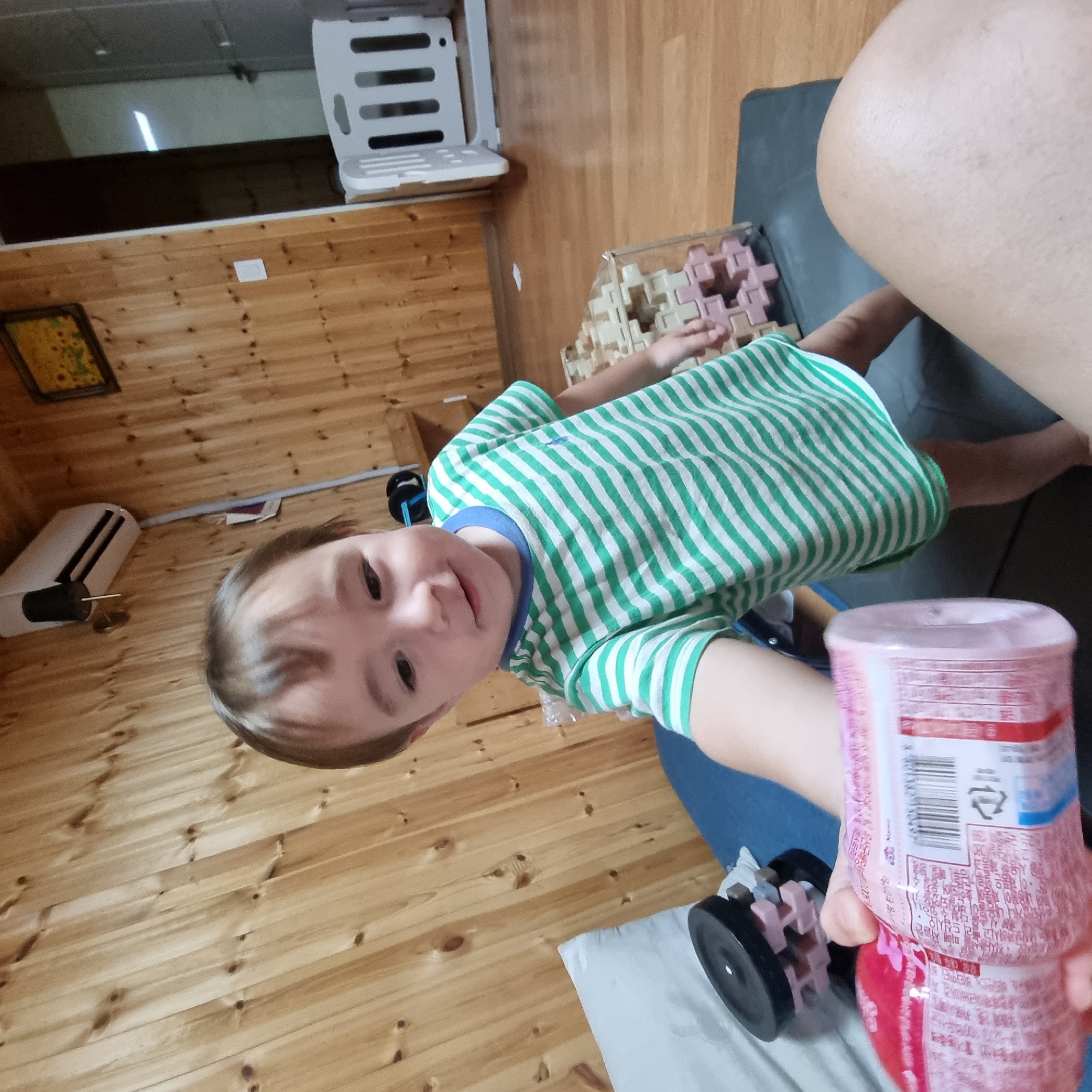 A young child, likely a toddler, wearing a green and white striped t-shirt, reaches out with their left hand holding a pink bottle. The child is positioned in the foreground, facing slightly to the left, with their head tilted down. Behind the child, in the middle ground, is a clear plastic container filled with interlocking, pastel-colored building blocks, some of which are pink and beige. To the left of the child, a dark object, possibly a weight, rests on a blue surface, which is likely a cushion or mat. The background features a wall paneled with light-colored wood, a white air conditioning unit mounted high on the wall, and a framed picture depicting sunflowers. Further in the background, a white baby gate is visible, suggesting a residential setting. The floor is a light wood. The scene appears to be indoors, within a home, and the image is taken from a low angle, suggesting the photographer is either also a child or positioned at a similar height. The location is identified as Dunnae-myeon, South Korea.