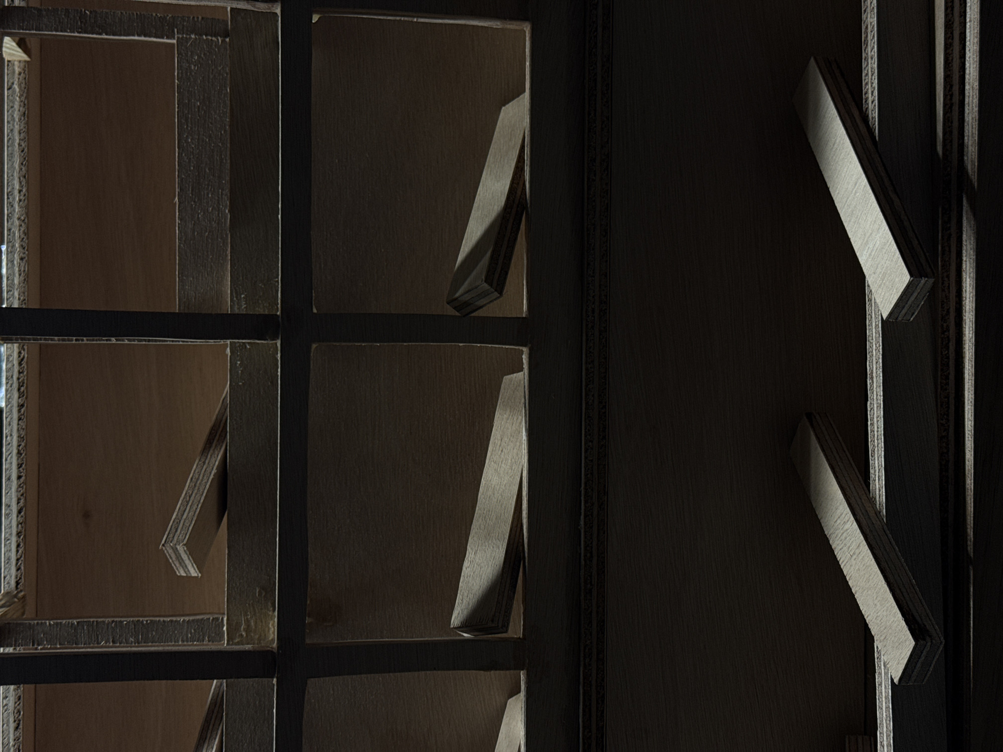 The image depicts a close-up view of a structure made of wood, possibly plywood or a similar composite material. It features a grid-like pattern with rectangular openings. Within these openings, smaller wooden pieces are positioned at angles, creating a sense of depth and shadow play. The lighting is dim and directional, emphasizing the textures of the wood and casting strong shadows. The color palette is muted, primarily consisting of browns, grays, and blacks.
Given the prevalence of steel and industrial history in Esch-sur-Alzette, Luxembourg, this structure could potentially be an artistic installation or architectural detail within a repurposed industrial building or a modern construction drawing inspiration from the area's industrial heritage. It is difficult to determine the exact location or purpose of the structure without further information.
