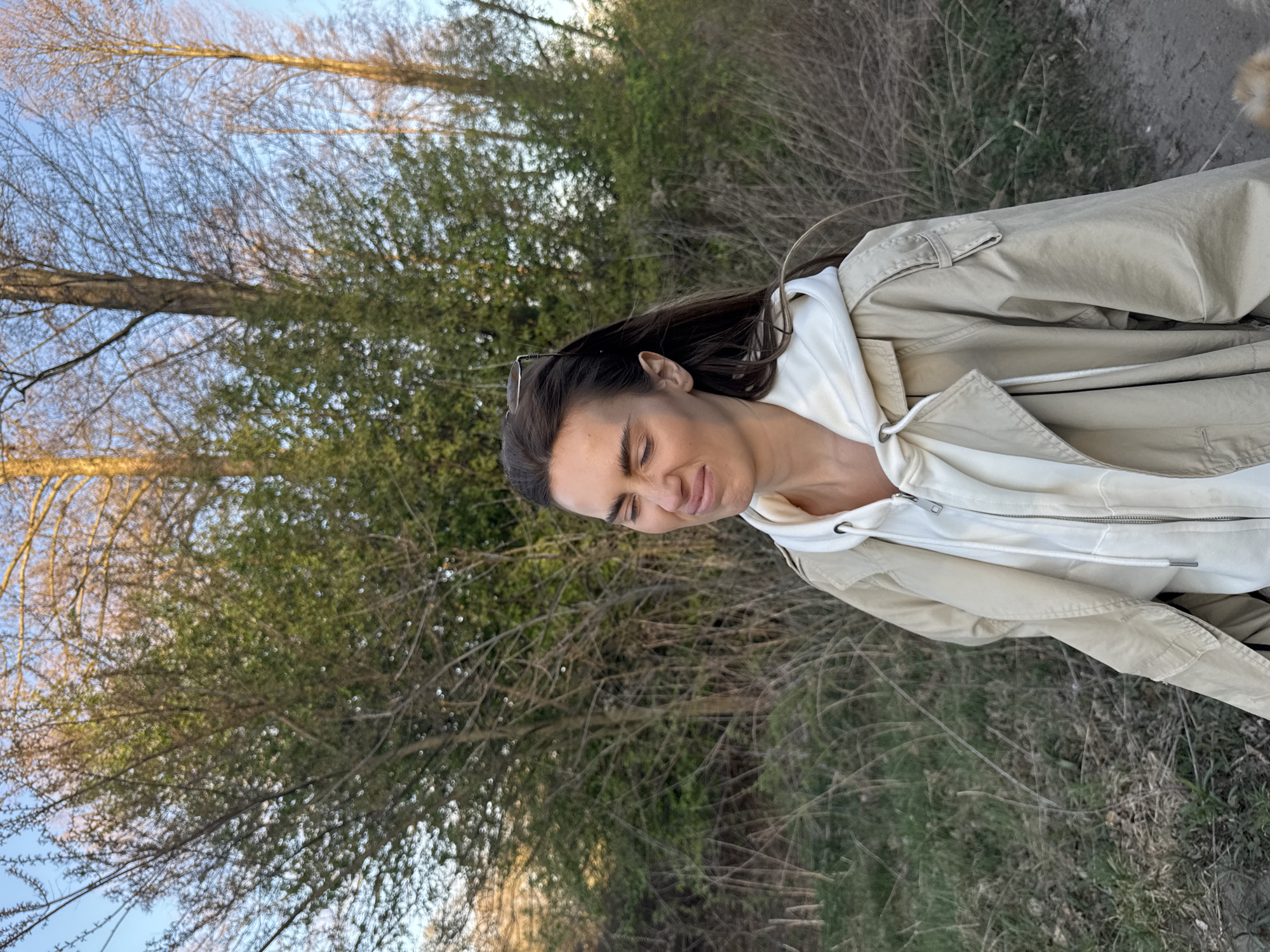 The image shows a woman outdoors, likely in a park or wooded area. She is wearing a light-colored hoodie under a tan jacket. The background features trees with bare branches and some green foliage. The ground is covered in grass and dry vegetation. The image is slightly rotated. The city of Arciechów, Poland, is not discernible from the image.