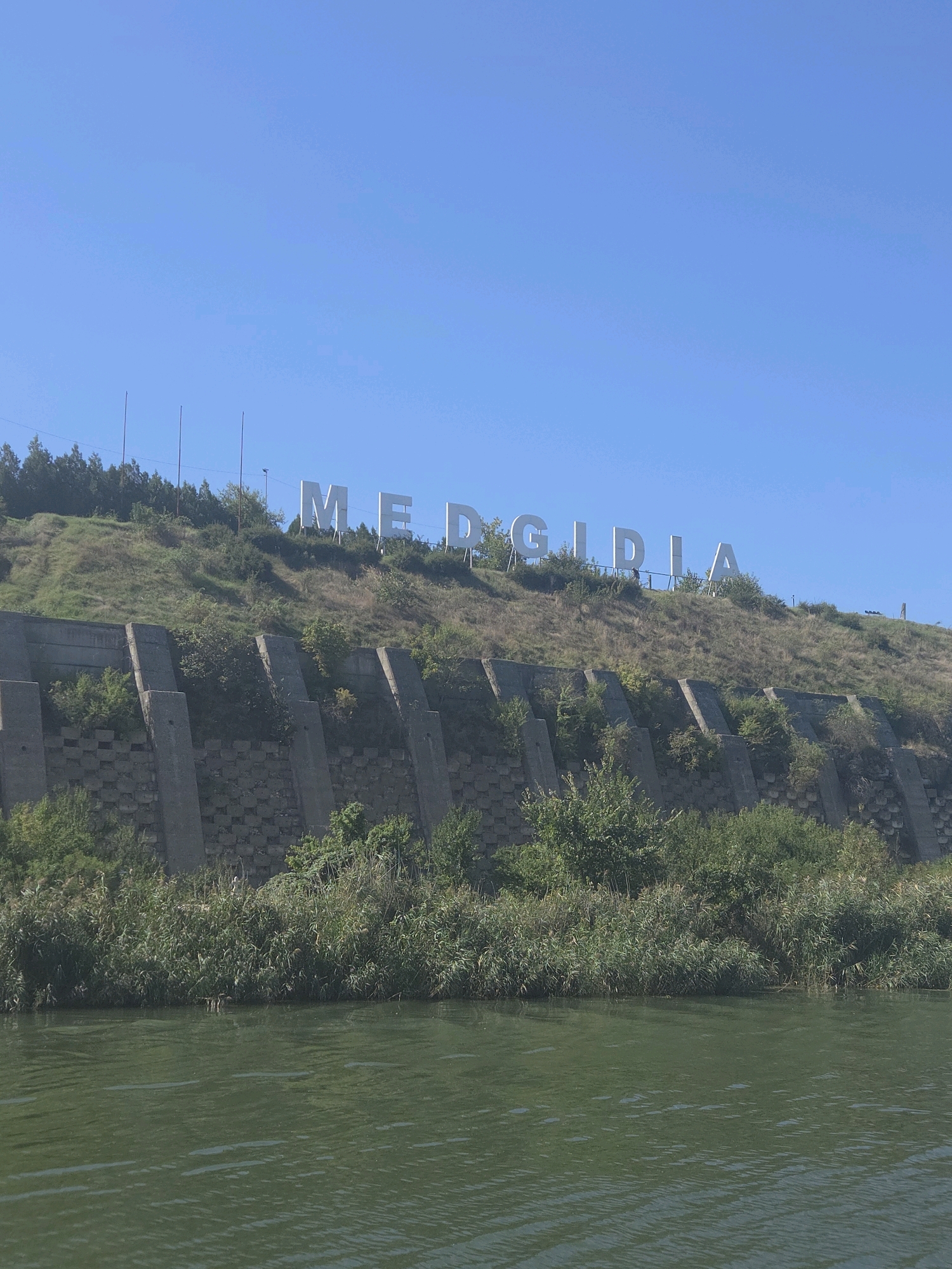 The image depicts a view from the Danube-Black Sea Canal, looking towards a hillside in Medgidia, Romania. Dominating the hillside are large, white, three-dimensional letters spelling out "MEDGIDIA." The hillside itself is covered in green grass and scattered trees, with a visible retaining wall constructed of concrete elements at its base. The canal water in the foreground is a deep green, with ripples on its surface. The sky above is a clear, bright blue. The overall scene is a landmark sign for the city, viewed from the waterway that passes by.