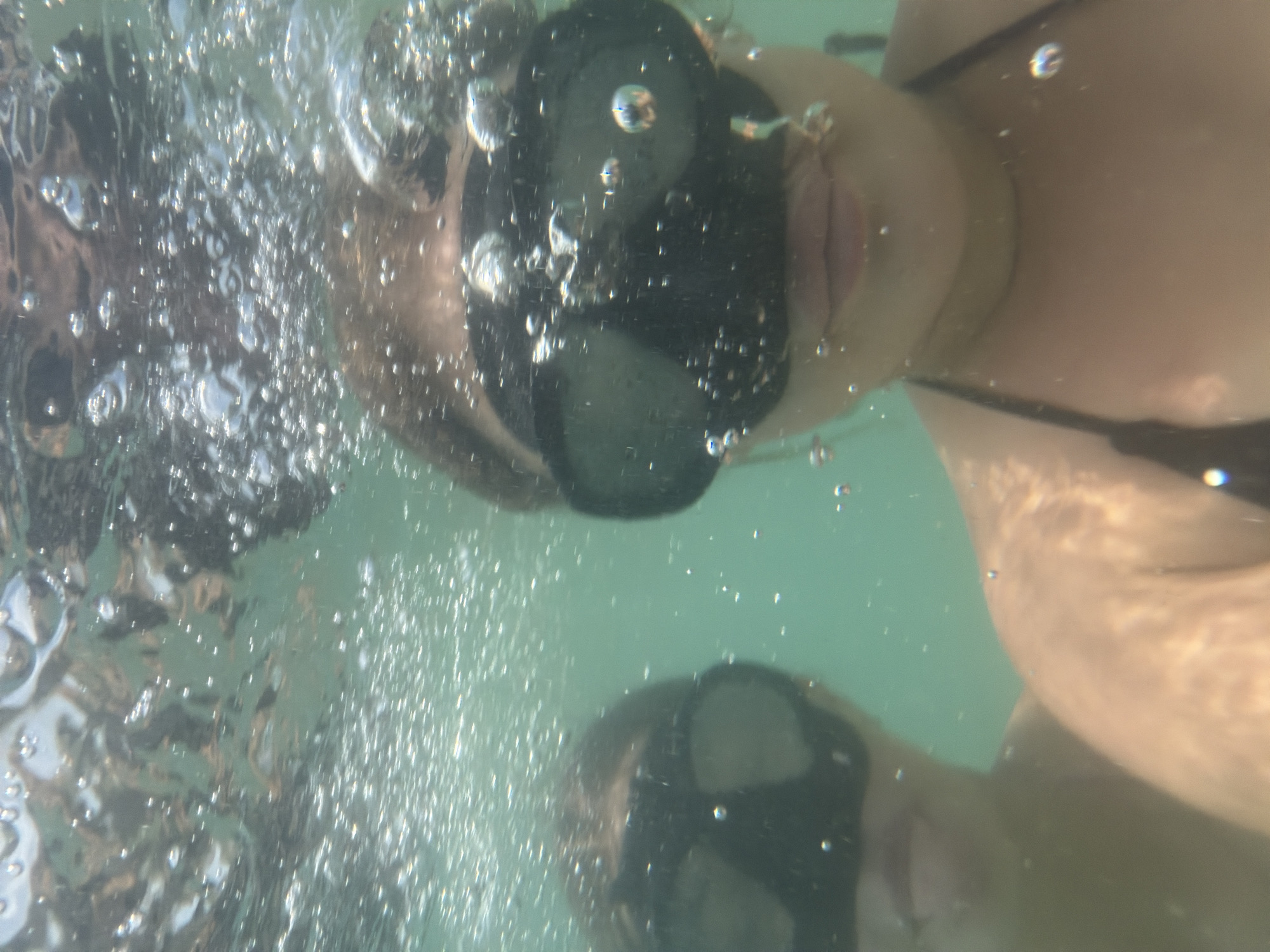 Two individuals, appearing to be young adults, are underwater, posing for a selfie. Both are wearing snorkeling masks, obscuring their eyes. The person in the foreground, on the right, is positioned closer to the camera and has their face mostly visible, showing their lips and part of their chin. They are wearing a dark-colored bikini top. Behind and to the left of this person, another individual's face is partially visible, also wearing a snorkeling mask. Numerous air bubbles are present throughout the water, with some clustered around the faces and others floating near the surface. The water is murky green, with light filtering from above, creating a slightly distorted view. The background is a blur of underwater elements, including reflections and shadows. The scene appears to be a recreational activity in a natural body of water, likely near a coastal area. The image does not provide specific indicators of the city of Nha Trang, Vietnam, beyond the general context of a tropical or subtropical marine environment.