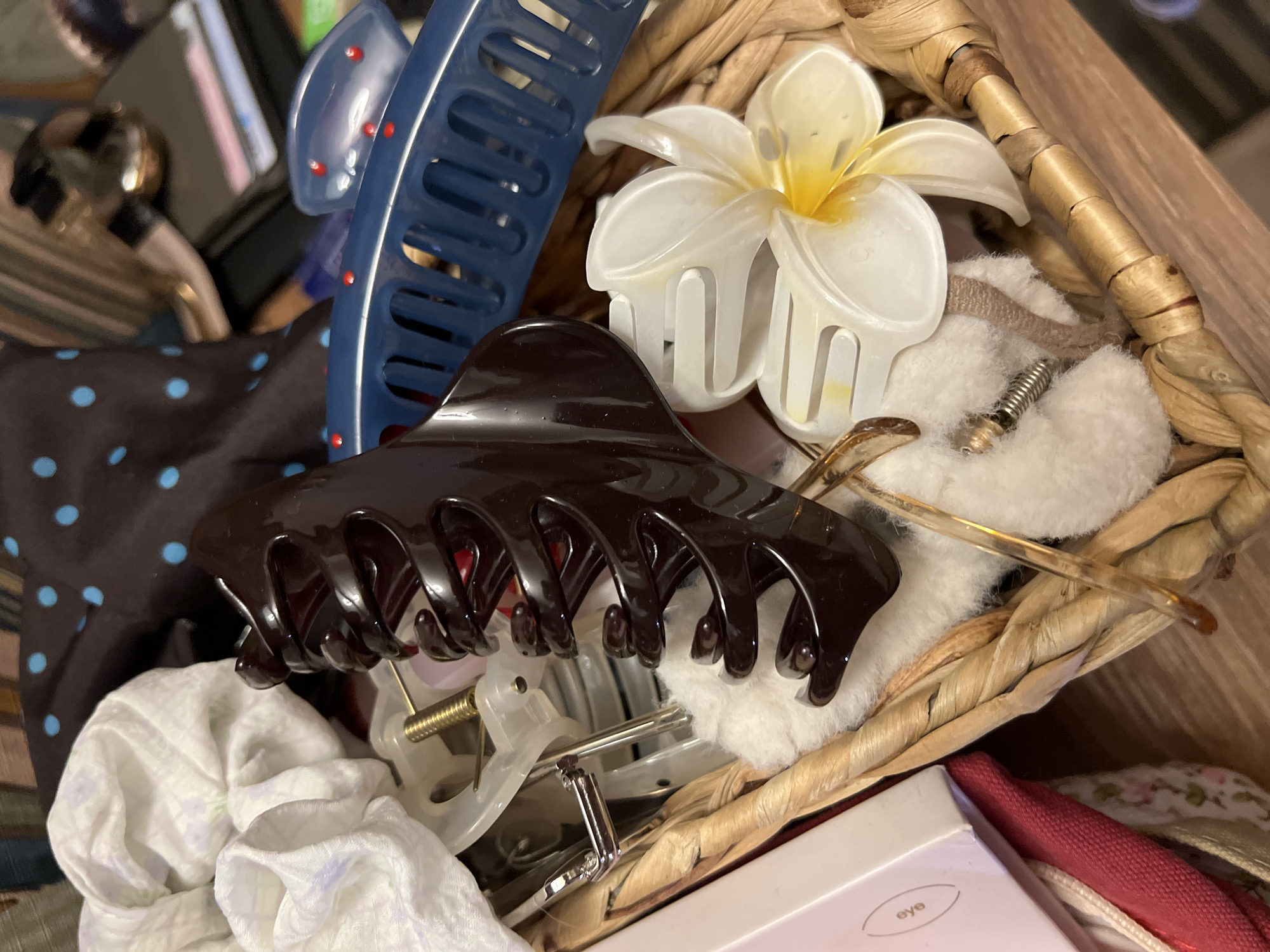 The image shows a close-up of a woven basket filled with various personal items, predominantly hair accessories and decorative elements. A large, dark brown plastic hair claw is prominently positioned in the center, its teeth open. To the right of the claw, a white plastic hair clip shaped like a plumeria flower with yellow accents is visible. Behind the claw, a blue plastic comb with red dots is seen. In the upper left, a crumpled white fabric with a faint floral pattern is present. Other items in the basket include a small metal spring, a thin pair of eyeglasses with translucent frames, and what appears to be a small pink box with the word "eye" printed on it. The overall impression is one of a collection of hair styling and beauty accessories, possibly stored in a drawer or on a vanity. The background is blurred, but suggests other objects and textiles, including a dark fabric with blue polka dots. There are no people or animals visible, and no discernible urban or natural environment. The image provides no information to suggest a location in the city of Unknown, Russia.