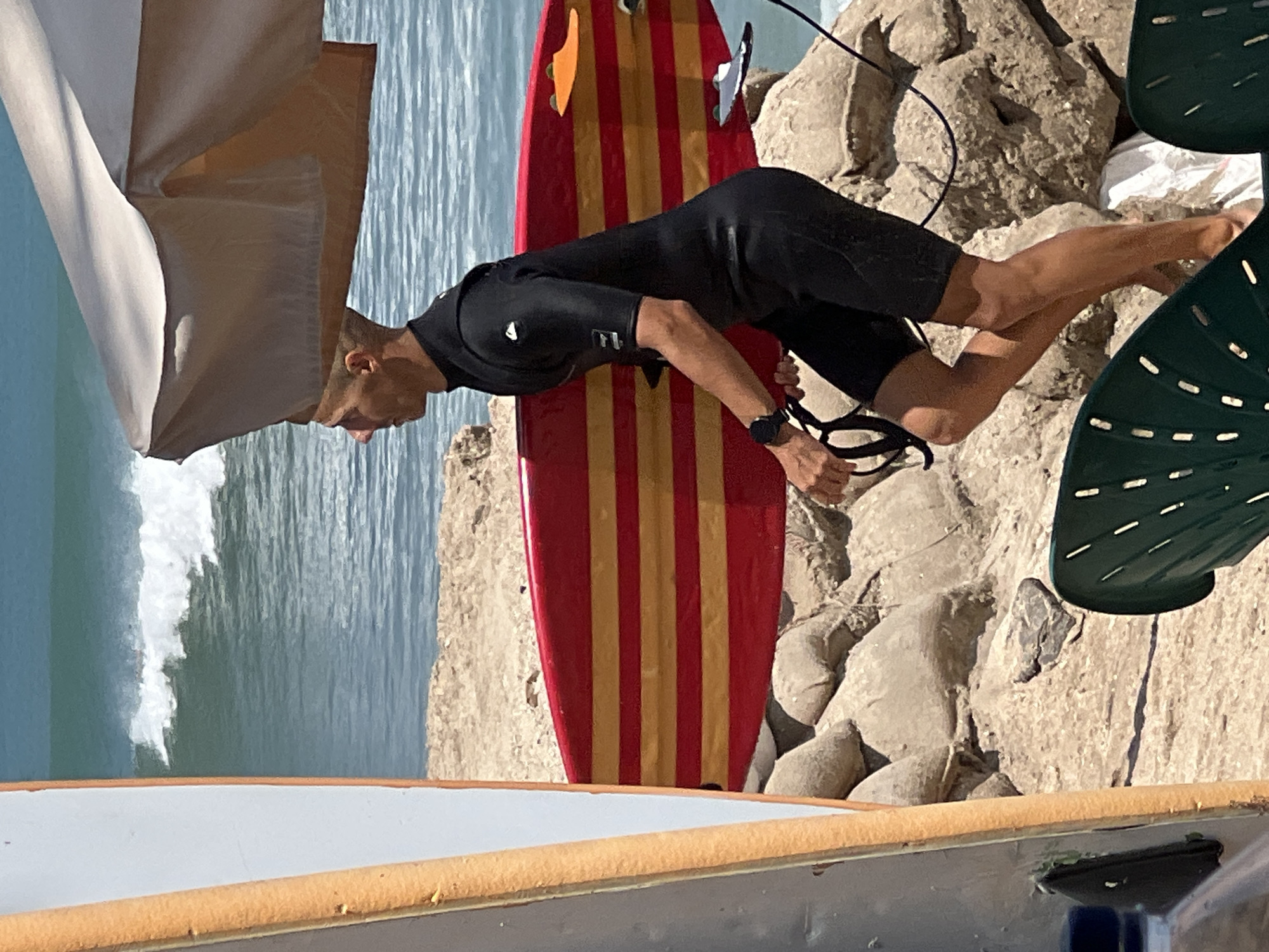 A surfer wearing a black wetsuit and black shorts walks along a rocky beach in Kuta, Indonesia, carrying a red and yellow striped surfboard. The surfer is seen from the side, with their head slightly down. They are holding the surfboard with their right hand, and a leash in their left hand. In the background, there is a wavy ocean with a breaking wave. The beach is made up of sand and rocks. There is a partially visible surf board to the left of the frame and a part of a beach umbrella above the surfer. Green chairs are visible in the foreground.