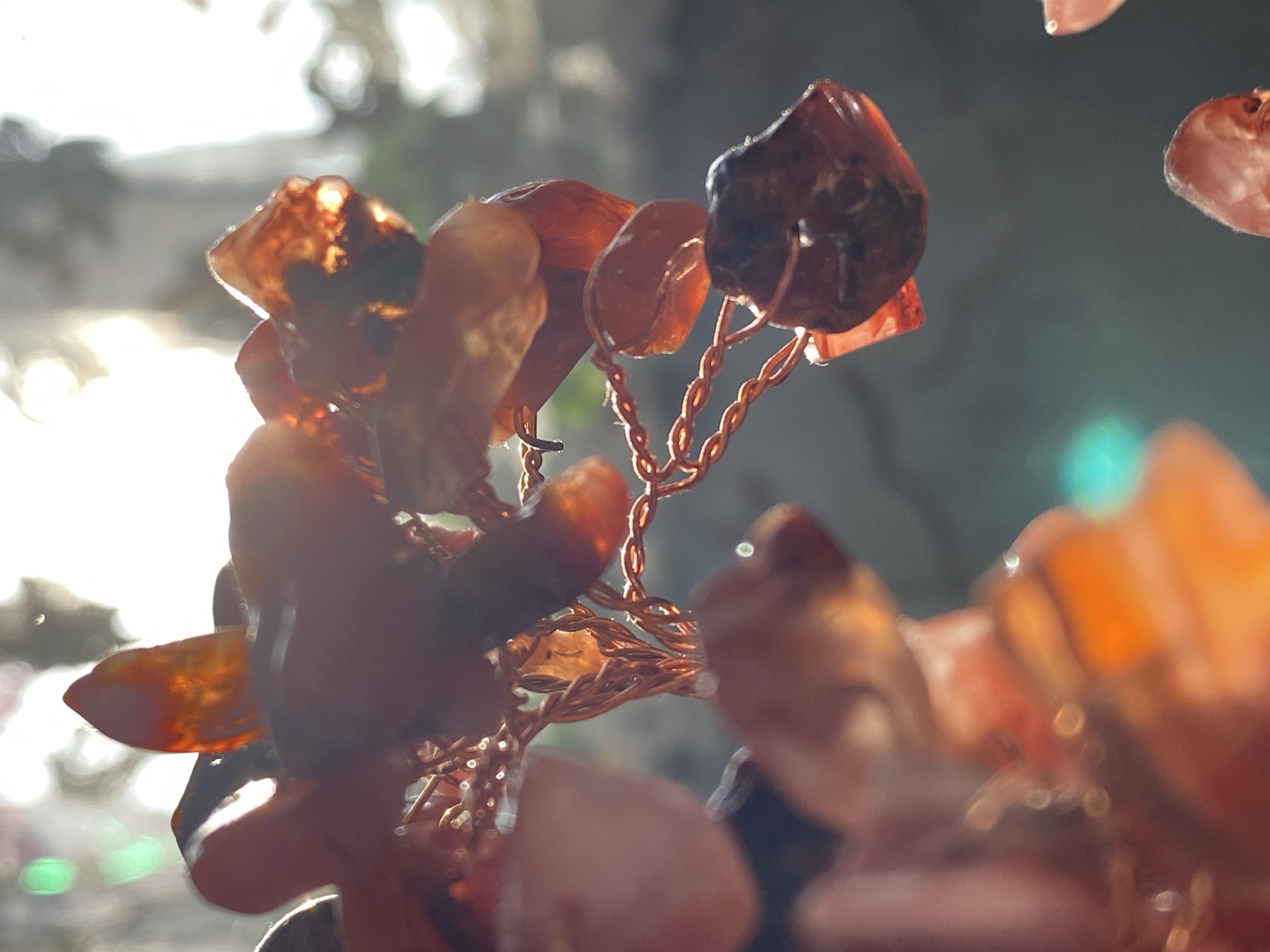 This close-up image features a decorative object composed of numerous small, irregularly shaped, translucent reddish-orange stones or beads, each individually wired with twisted copper strands. The stones resemble carnelian or similar semi-precious gems, some appearing deep amber and others glowing brightly as light passes through them.
The object is strongly backlit, likely by natural light from a window or the sun, creating a high-key and atmospheric scene. The background is heavily blurred (bokeh), characterized by bright, amorphous white and yellow light on the left, indicating the dominant light source. Darker, indistinct shapes with subtle green hues can be observed in the background on the right. The strong backlighting accentuates the translucency of the stones and the intricate details of the copper wire, which forms delicate branches or connections.
No activity or event is taking place; the image is a static portrayal of the decorative item. The time of day is suggested to be bright, possibly morning or late afternoon, given the warm tones and dramatic lighting. There are no specific location cues visible within the image itself. No text is visible. The provided location context is Фоки, Russia.