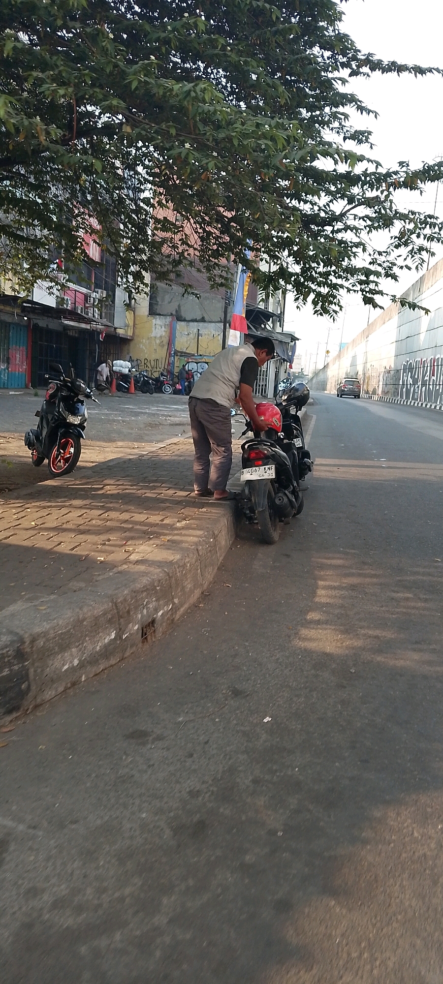 A man in a grey vest and grey pants is seen from the side, standing on a sidewalk next to a black motorcycle in Bekasi, Indonesia. He is bending down, looking at the motorcycle and holding a red object, possibly a helmet, in his hands. Another black motorcycle is parked to his left. In the background, there are buildings with signs and a street with a few other motorcycles parked. A car is visible further down the street, and a concrete wall with graffiti runs along the right side of the road. The scene appears to be in an urban environment, with a tree overhead casting shadows.