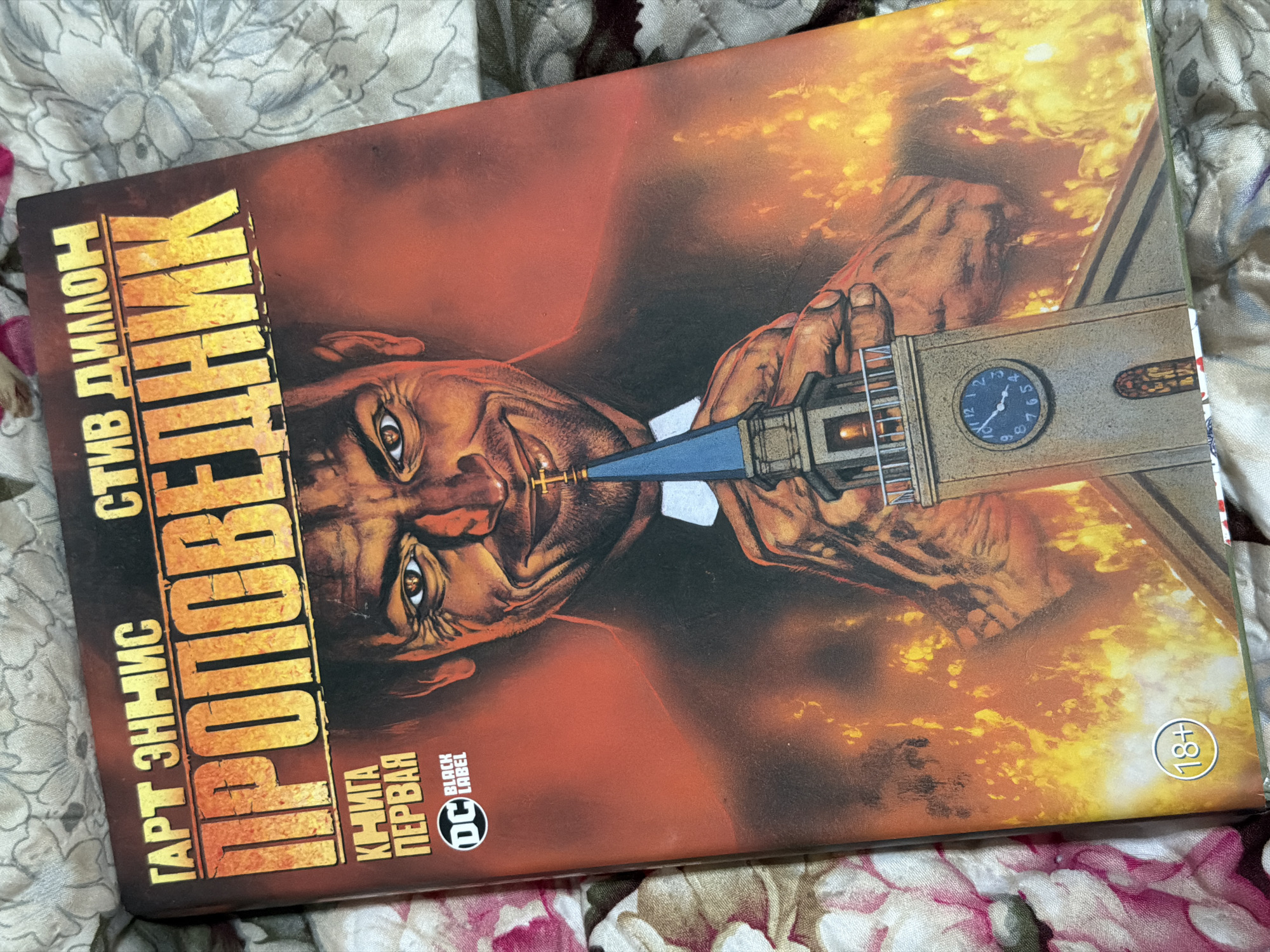 This is a close-up, top-down photograph of the cover of a comic book. The comic book cover features a dark, fiery background with an illustration of a man in a clerical collar and suit, holding a church steeple with a clock face. The man has a sinister smile and his eyes are intensely lit. The title of the comic book, in large, distressed yellow lettering, reads "ПРОПОВЕДНИК" (Propovednik), which translates to "The Preacher". Above the title, in smaller yellow lettering, are the names "ГАРТ ЗННИС" (Garth Ennis) and "СТИВ ДИЛЛОН" (Steve Dillon). To the left of the title, in smaller orange lettering, it says "КНИГА ПЕРВАЯ" (Book One) and includes the DC Black Label logo. In the bottom left corner of the cover, there is a circular "18+" rating symbol. The comic book is lying on a textured surface, possibly a blanket or fabric with a floral pattern in shades of pink, white, and gray. There is no indication of the specific location in Moscow, Russia, or any other city.
