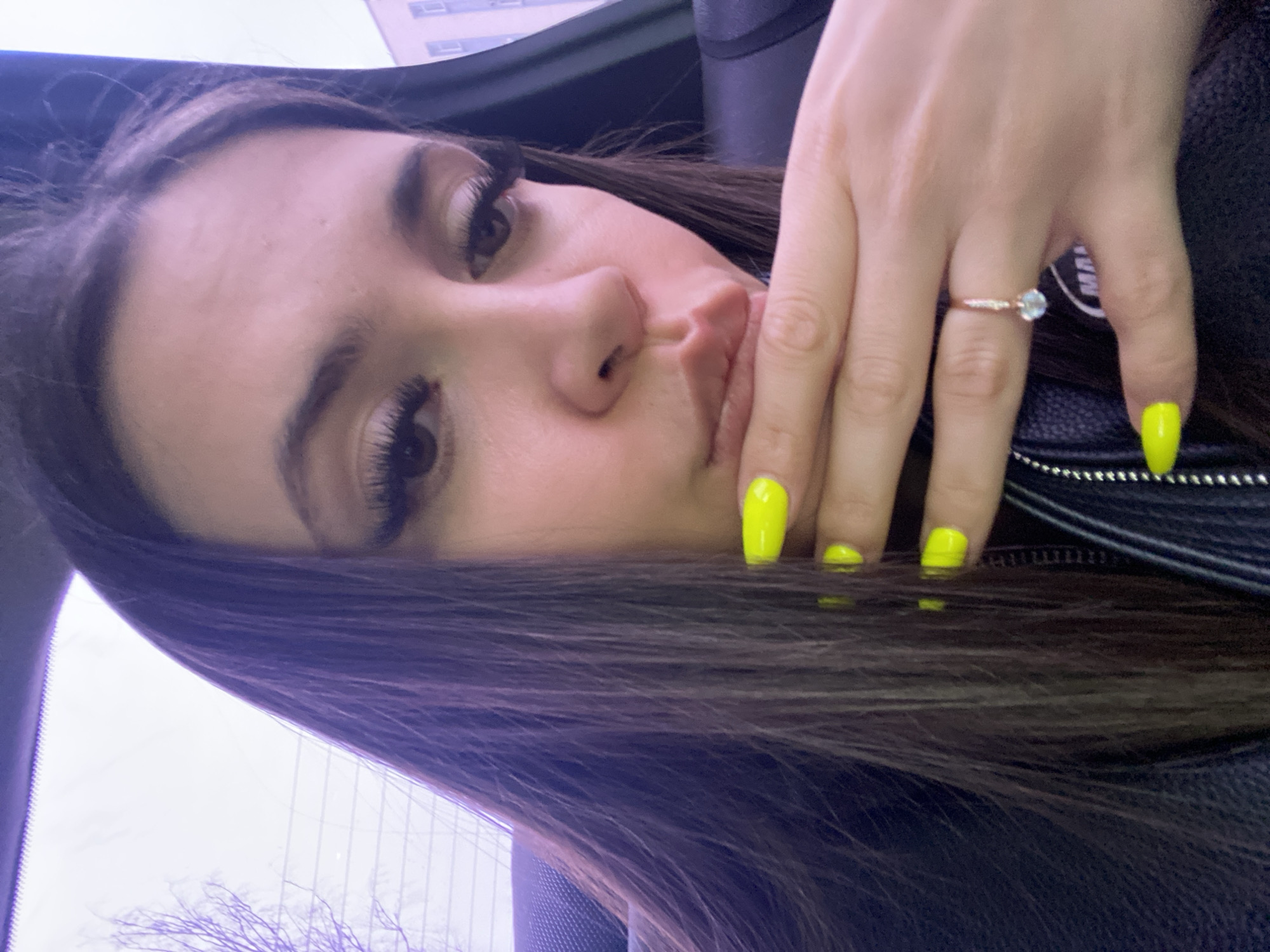 A selfie taken in an enclosed space, possibly a car, shows a young woman with long, dark brown hair and fair skin. She is wearing dark clothing, possibly a leather jacket. Her left hand is partially obscuring her face, revealing bright yellow nail polish. A ring with a light-colored gemstone is visible on her ring finger. In the background, through a window, a blurred cityscape with a building is visible, suggesting an urban setting. The presence of bare tree branches outside the window hints at either late autumn or early spring. Based on the request for the city of Chaikovsky, Russia, the scene is likely set there, though there's no explicit confirmation from the visual content.