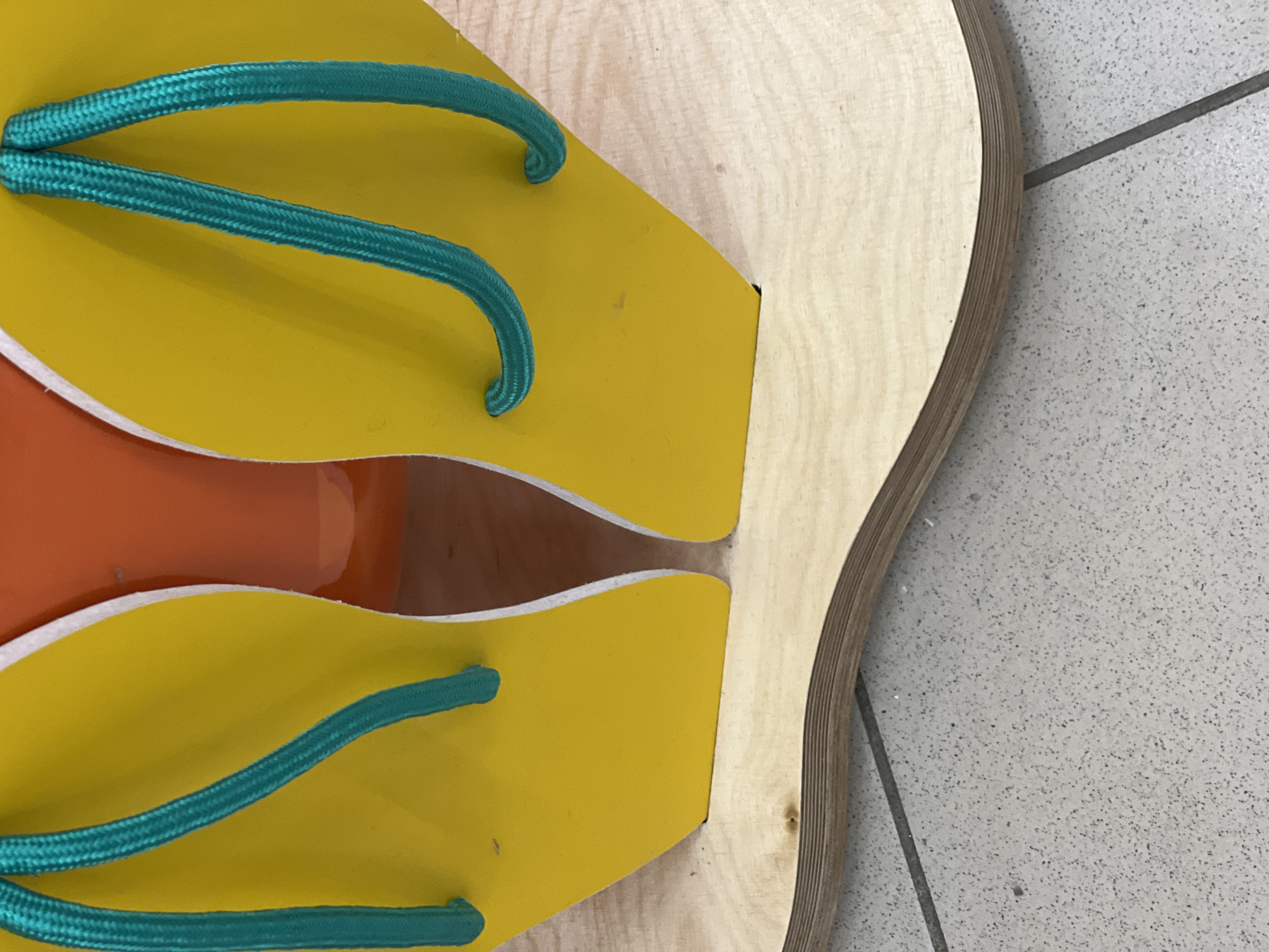A close-up, overhead shot reveals a pair of oversized, yellow flip-flops with teal straps resting on a light-colored, curved wooden platform. The flip-flops have white edging. Behind the flip-flops, an out-of-focus orange object is visible. The wooden platform sits on a grey tiled floor. The scene does not provide any specific information to place it in Yekaterinburg, Russia.