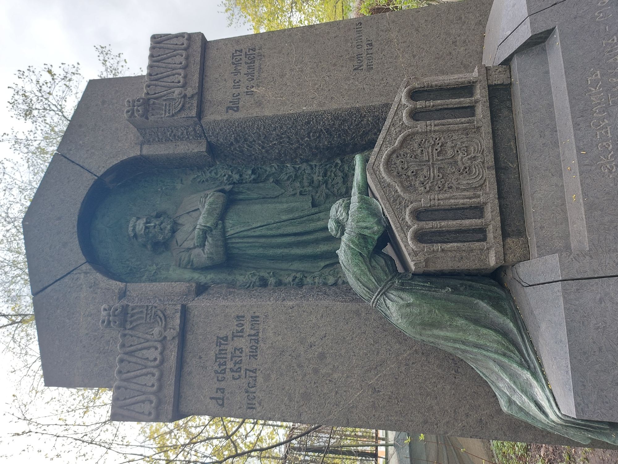 This is a vertical, medium-shot photograph of a large, gray stone monument. The monument has a bas-relief sculpture of a bearded man in religious robes, set in an arched alcove. The sculpture is green in color. To the right of the man is a three-dimensional sculpture of a female figure, also green in color, kneeling and reaching towards a stylized book or ornate screen. Text in Cyrillic is carved into the stone on either side of the man's figure. Latin text is also present at the top right of the stone. A cityscape with trees is visible in the background. This image is likely of a statue in a cemetery in Saint Petersburg, Russia.