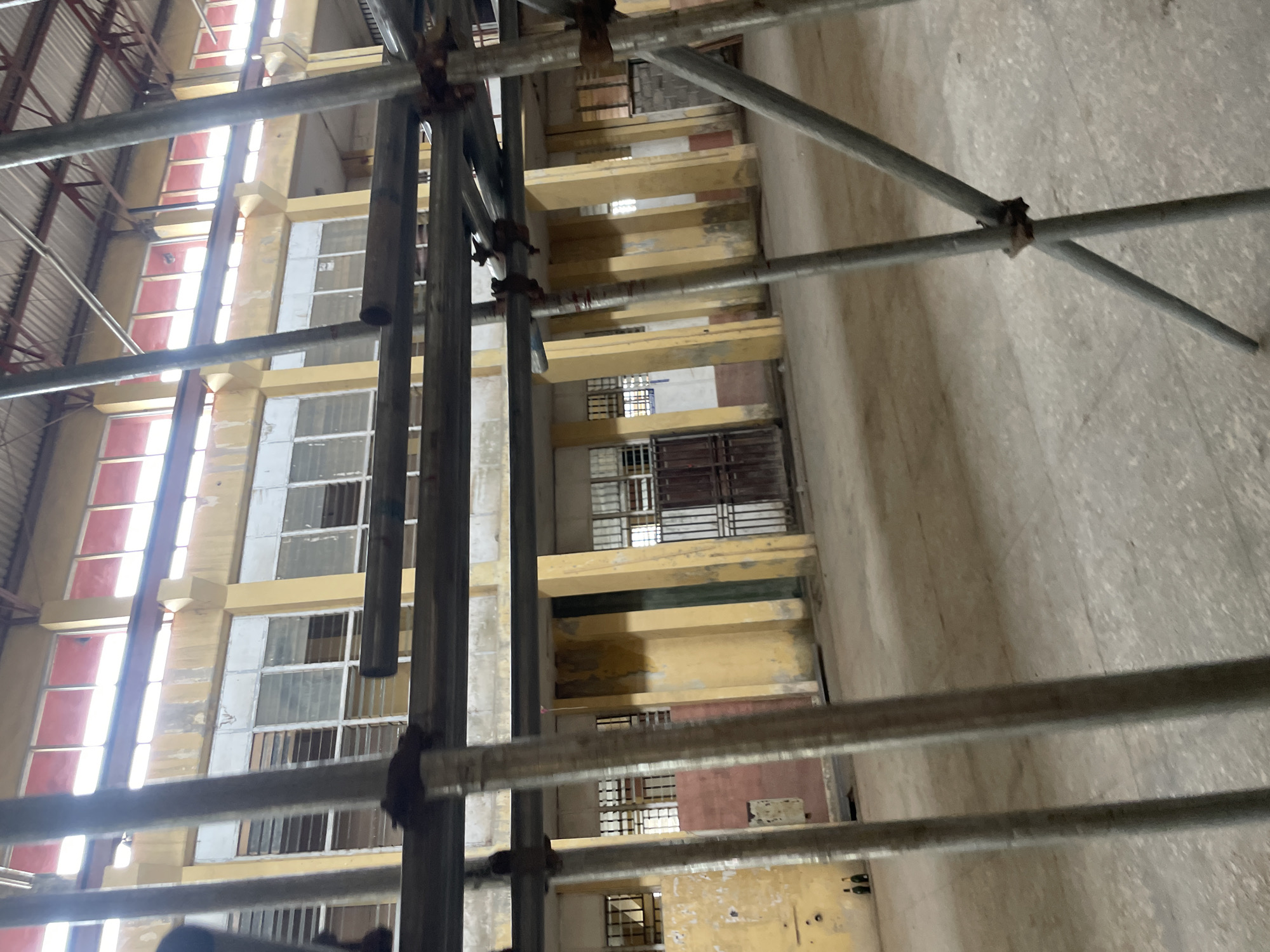 The image shows a large, empty indoor space that appears to be under construction or renovation. Scaffolding made of metal pipes is prominently featured in the foreground, obstructing a clear view of the environment. The walls are painted a pale yellow, with some areas showing discoloration or peeling paint. There are numerous windows along the walls, many of which are covered with horizontal metal bars or louvers. The windows on the upper level have red frames. The floor is a light-colored concrete surface. In the background, there are doorways and some brickwork is visible. Two dark bottles are placed on the floor near the left wall. The overall impression is of a large, industrial or institutional building. The location is identified as Ihiagwa, Nigeria.