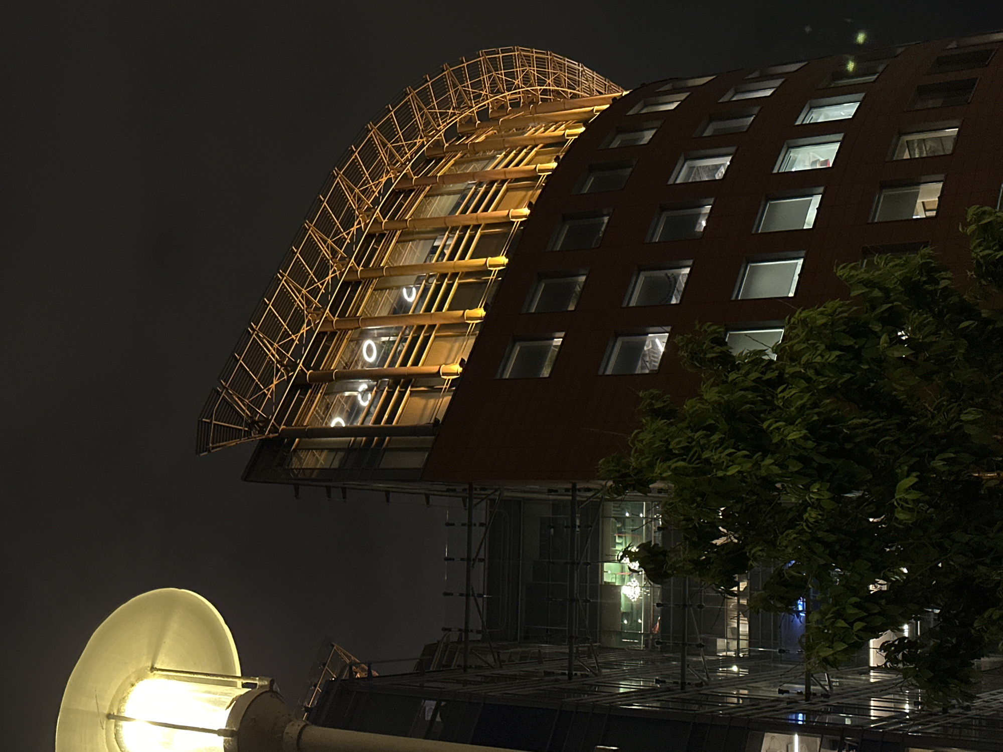 The image captures a nighttime scene in Moscow, Russia, focusing on a distinctive architectural structure. The building features a curved, external framework resembling scaffolding, contrasting with the building's facade, which has regularly spaced, illuminated windows. A street lamp illuminates the foreground, while a tree partially obscures the lower-right section of the building. The sky is dark, suggesting the photo was taken at night or during low-light conditions.