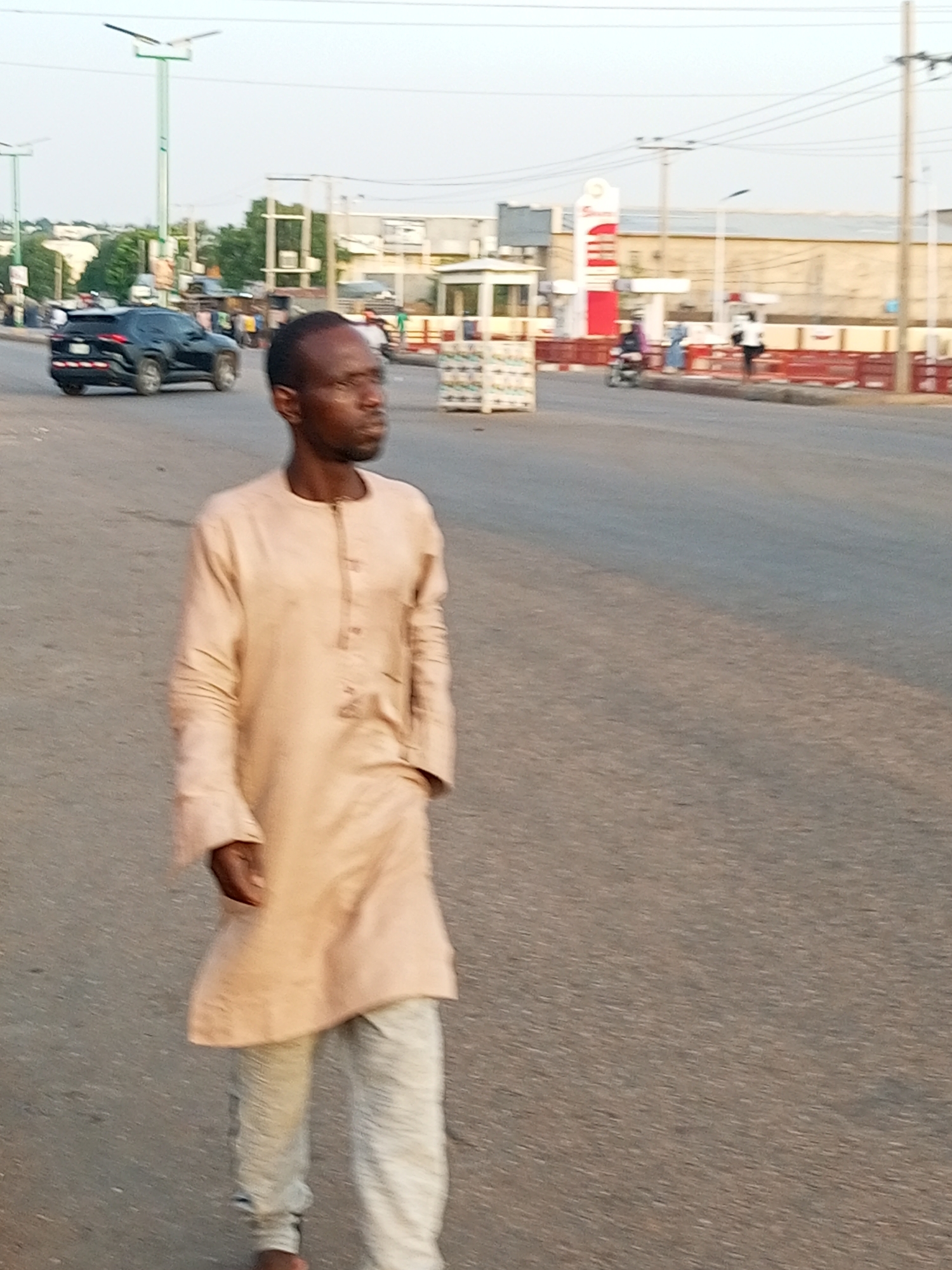 A man is walking on a street in an urban setting. He is wearing a long-sleeved, light-colored shirt and pants. A black car and other vehicles are visible on the road. A gas station is in the background, along with other buildings and structures. There are power lines above. The location appears to be an intersection or a busy area in the city.