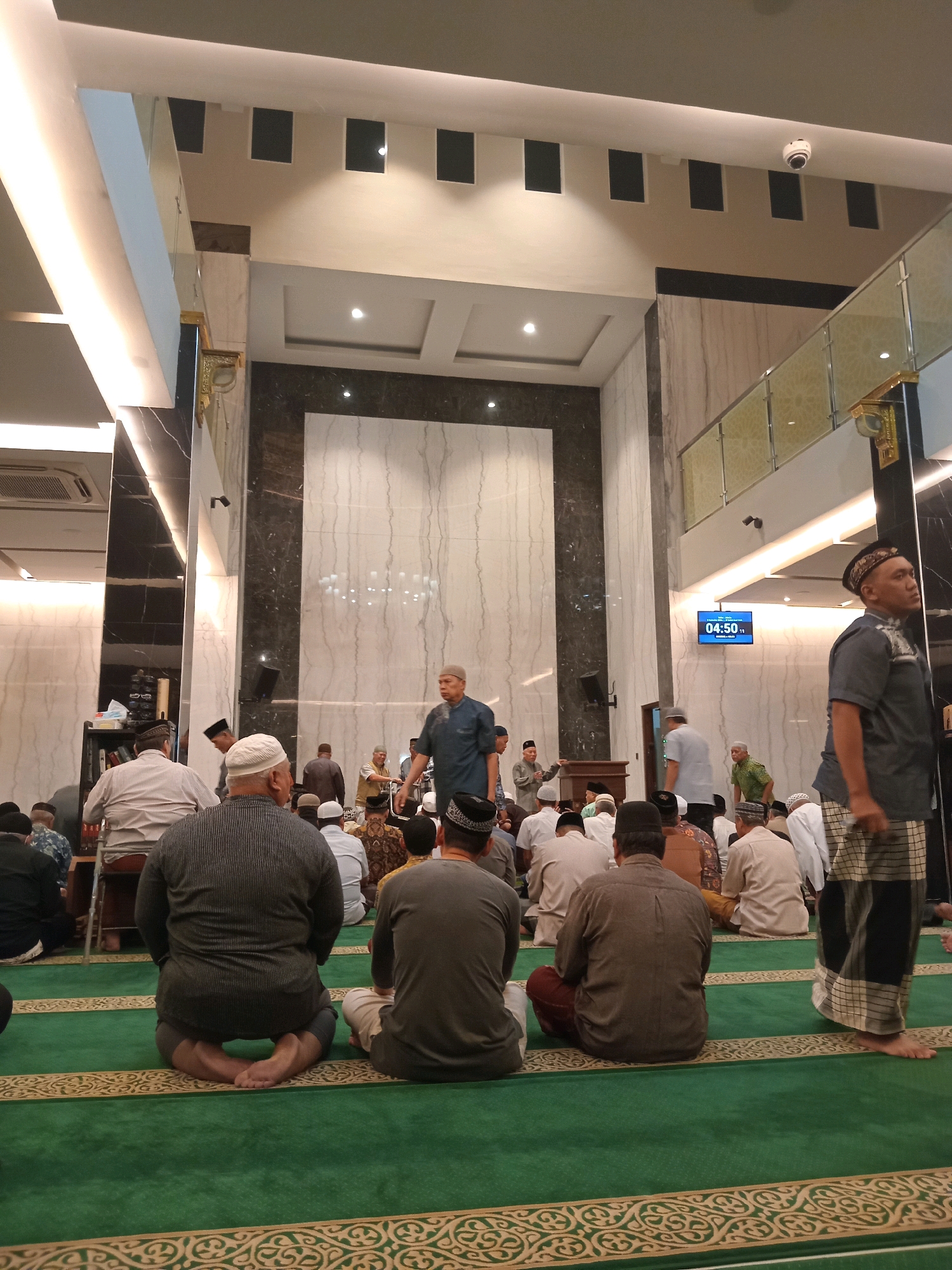 This image depicts a gathering of approximately 30 Muslim men inside a mosque, likely for prayer. The men are seated on a green carpeted floor with ornate gold trim. The architecture of the mosque is modern, with high ceilings, marble walls, and recessed lighting. In the background, a digital clock displays "04:50". A few individuals are standing, and one man in a blue shirt appears to be speaking or leading the group. A security camera is visible on the ceiling. The overall atmosphere suggests a religious service or communal prayer session.