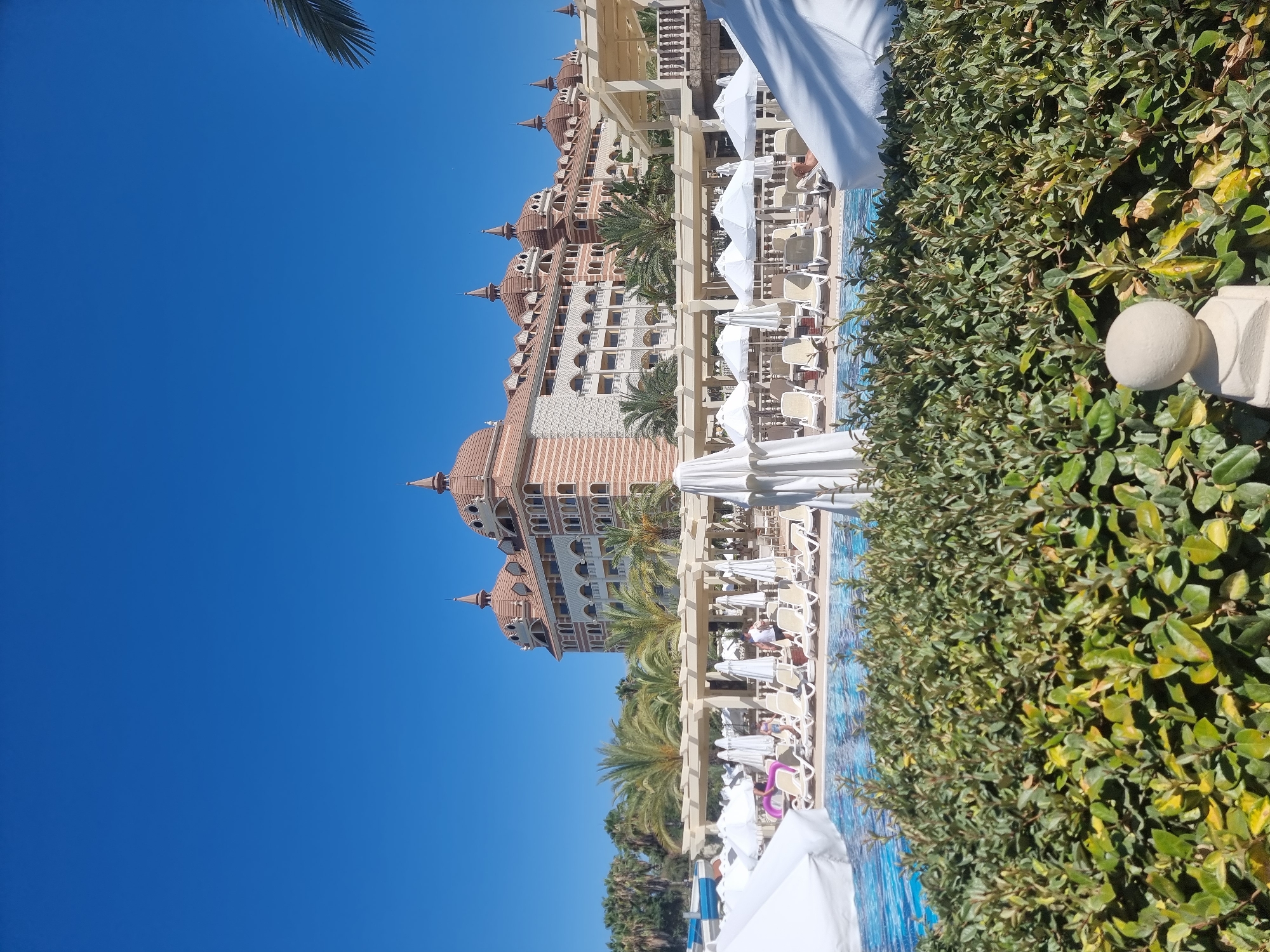 In Manavgat, Turkey, a bright, sunny day is captured in this image of a resort with a swimming pool and a large hotel. The hotel, with its distinctive domes and patterned facade, stands tall against a clear blue sky. In the foreground, a swimming pool with blue water is visible, surrounded by rows of empty lounge chairs and white umbrellas. Lush green bushes line the edge of the pool area, and palm trees are scattered throughout the scene, indicating a tropical or subtropical setting. A few people are seen near the pool, some lounging in chairs and others walking around. The overall impression is one of a luxurious and relaxing vacation destination.