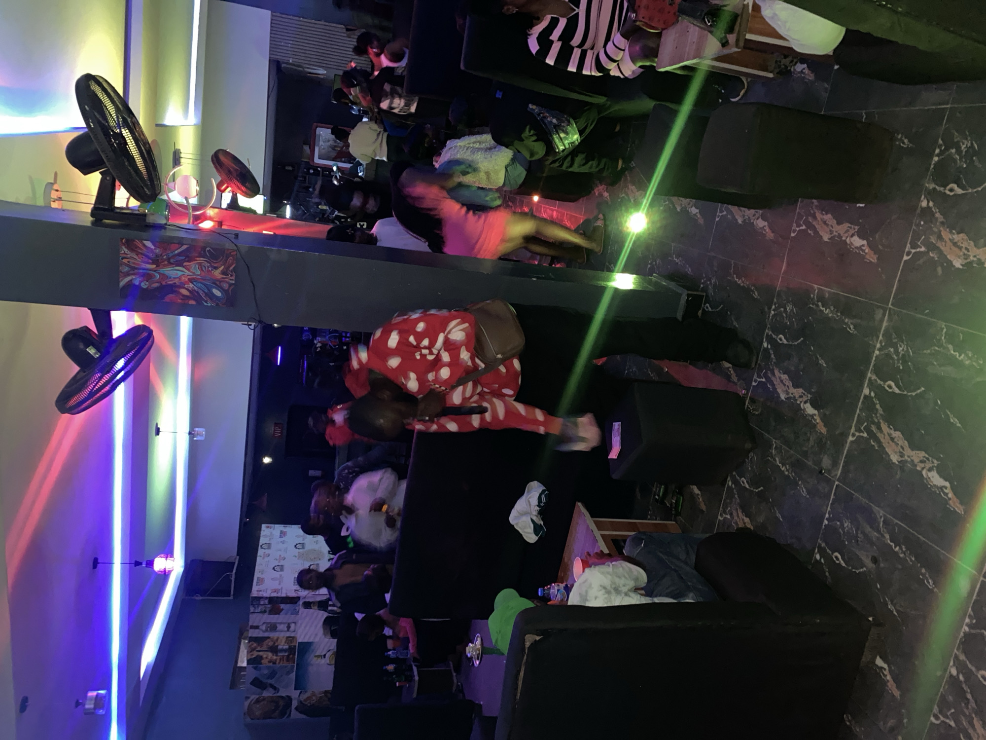 The image captures an indoor scene, possibly a nightclub or lounge, with multiple people present. The lighting is dynamic, with streaks of red, blue, and green light illuminating the space, suggesting the use of party lights. Several large ceiling fans are visible, some angled towards the ceiling, others pointing downwards. The floor is tiled with a dark, marble-like pattern.
In the foreground, a person in a red and white patterned outfit is holding a microphone and appears to be performing or speaking. To their left, a person wearing a green hat is seated, and a table with drinks and a cup is in front of them. To the right of the performer, a woman in a pink dress is interacting with another person. In the background, a group of people are gathered, some standing and others seated. There are black couches or seating areas arranged throughout the space. The walls appear to be painted in dark colors, and there are some decorative elements, including framed artwork. The overall atmosphere suggests a lively social gathering.
This scene is consistent with venues found in urban centers like Jos, Nigeria, where such entertainment establishments are common. The presence of multiple fans is a practical consideration in a climate like Nigeria's. The style of lighting and seating also aligns with typical nightclub decor in the region.