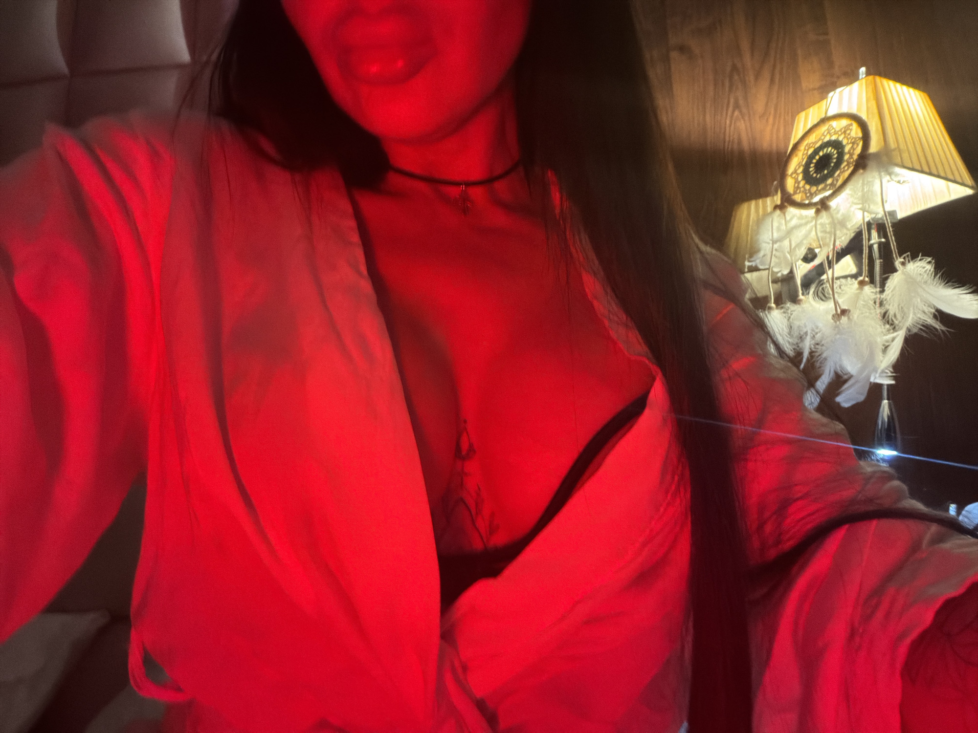 The image shows a person with long dark hair, wearing a light-colored robe or shirt that is open at the chest, revealing a bra. A thin black choker necklace is visible around the person's neck. The lighting is predominantly red. To the right of the person, there is a lamp with a lampshade and a dreamcatcher attached to it. The location appears to be indoors, possibly a bedroom. There is no verifiable information indicating the scene is in Boreti, Montenegro.