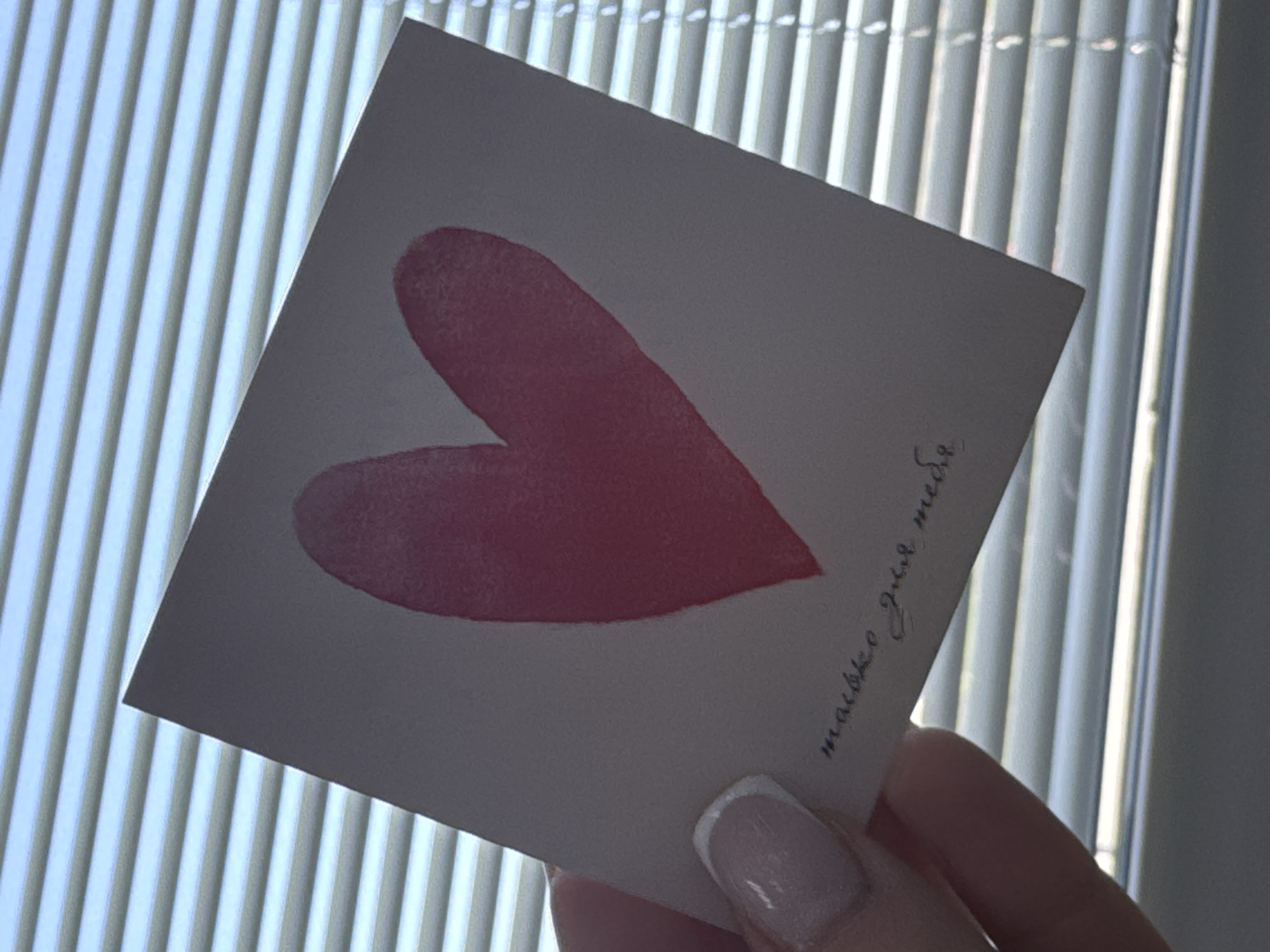 A slightly angled close-up shot depicts a person holding a greeting card in their fingers. The card, featuring a watercolor-style red heart, is held against a background of vertical blinds. Russian text "только для тебя" is written in cursive in the lower right corner of the card, translating to "only for you." The person's manicure is visible: a French tip style on their thumb. The background consists of window blinds and an exterior light source illuminating the scene. There is no information in the image that identifies a location in the city of Chaikovsky, Russia.