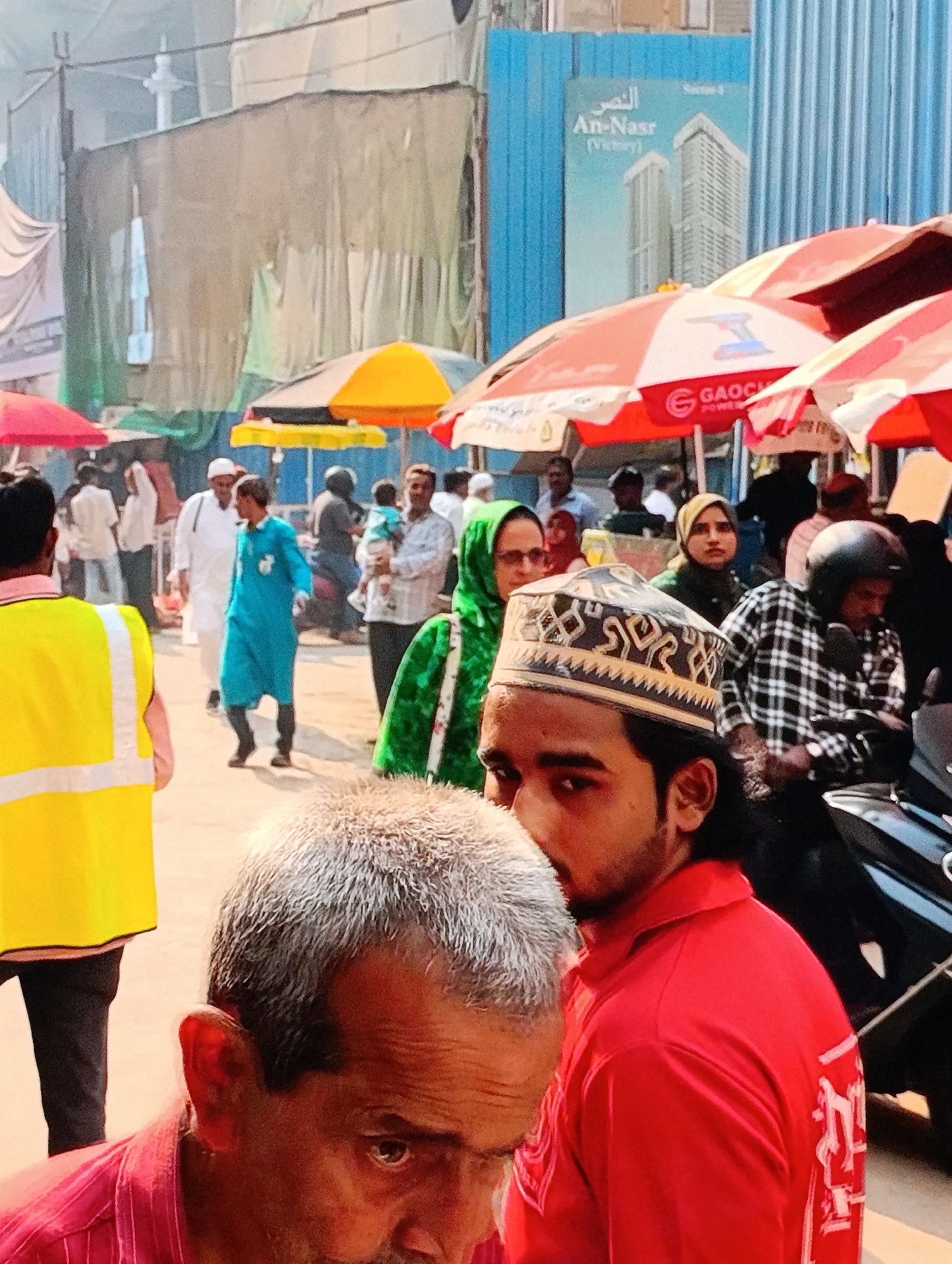 The image depicts a busy street scene in an urban location. Several people are visible, including men and women of varying ages, some wearing traditional Muslim attire. One man in the foreground wears a red shirt and a traditional Muslim cap with intricate patterns. Another man in a pink shirt is partially visible in the lower left corner, with gray hair. There is a person wearing a yellow reflective vest on the left side of the frame. Further back, a man is holding a baby.
Several umbrellas, predominantly red and white, provide shade over what appears to be street vendors or market stalls. In the background, there is a blue construction barrier with a poster featuring a building design labeled "An-Nasr (Victory)". The environment suggests a crowded, active marketplace or pedestrian thoroughfare.