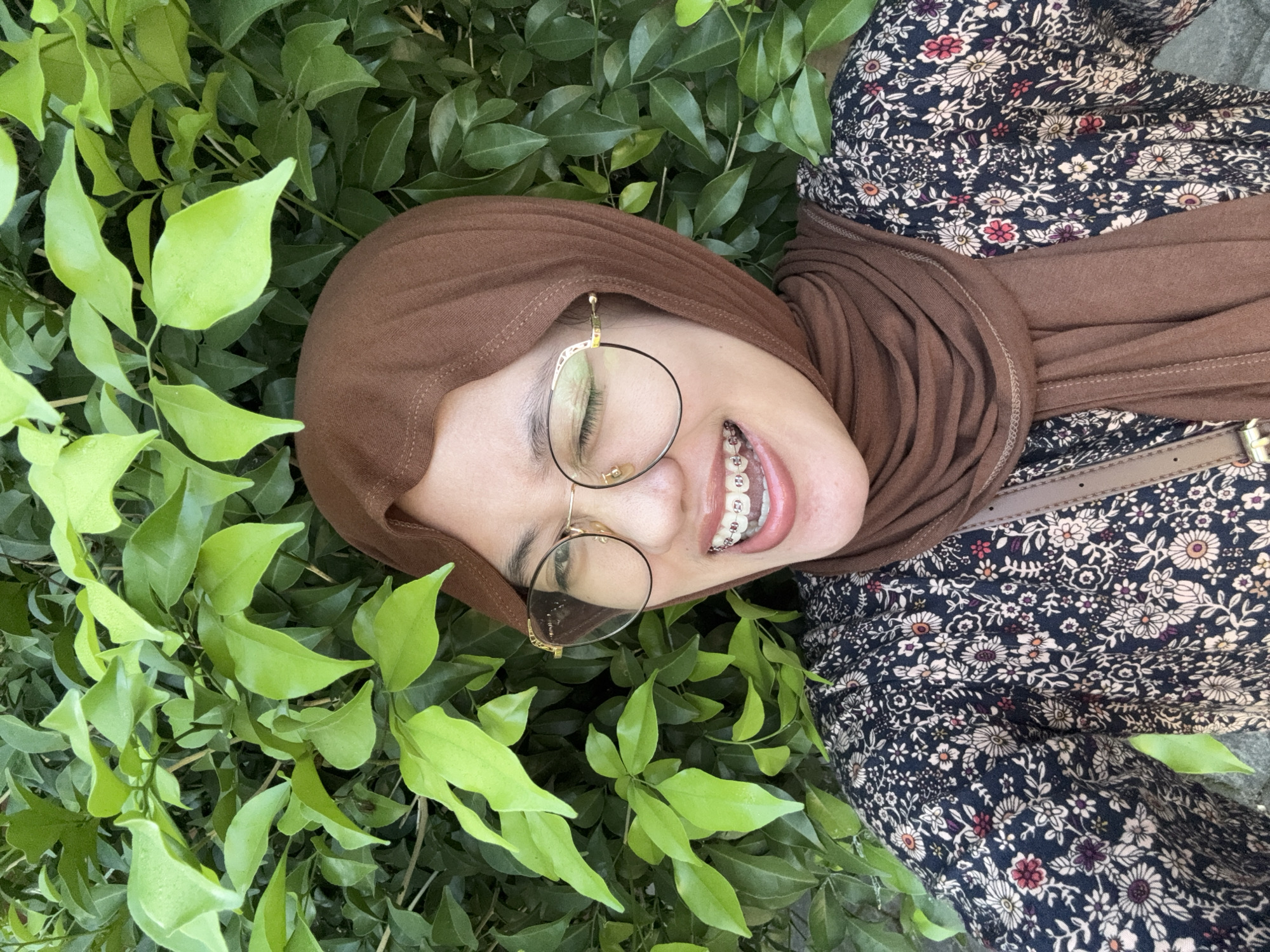 A young woman wearing a brown hijab, round eyeglasses, and braces is smiling with one eye closed, looking at the camera. She is wearing a dark, floral-patterned dress. A brown bag strap is visible across her chest. She is standing in front of a dense green hedge. The scene appears to be outdoors, possibly in a park or garden area in Gadingrejo, Indonesia, given the lush foliage.