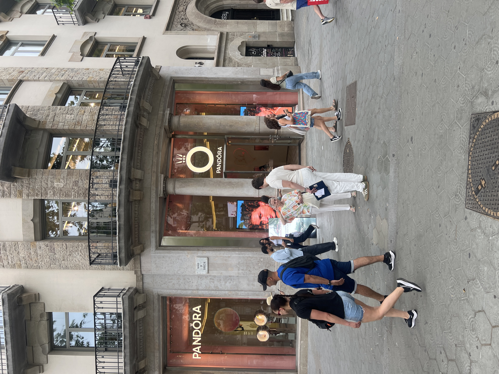 The image depicts a street scene in Barcelona, Spain, likely on Passeig de Gracia, indicated by a street sign visible on the building. The primary focus is on the facade of a Pandora jewelry store, characterized by its large glass windows displaying pink lighting and the Pandora logo. Several people are walking on the sidewalk in front of the store.
On the left, a woman in a black t-shirt and denim shorts walks past, followed by a man wearing a blue t-shirt, dark shorts, and a black baseball cap. To their right, a couple is walking away from the camera; the woman in front wears a white tank top and denim shorts, while the man behind her wears a light blue shirt and dark pants. Further right, a woman in a white top and floral skirt walks with a man in a white t-shirt and dark pants. On the far right, a person wearing a red shirt and dark shorts is partially visible, walking away from the camera.
The architecture of the building features stone accents, balconies with wrought-iron railings, and large arched doorways. The sidewalk is paved with hexagonal-shaped tiles. The overall atmosphere suggests a busy urban environment.