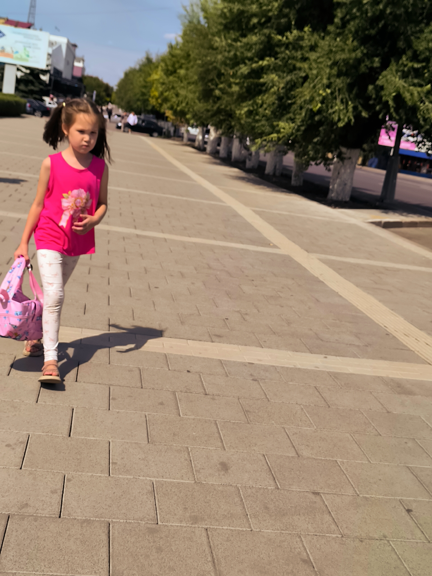 A young girl with dark hair in pigtails, wearing a bright pink sleeveless top and white patterned leggings, walks along a paved sidewalk. She carries a pink patterned bag over her left shoulder. The sidewalk is made of rectangular grey pavers with lighter colored borders. Trees line the right side of the sidewalk, with their trunks painted white. In the background, buildings and cars are visible, suggesting an urban environment. The sky is blue with some clouds. The image appears to be taken in Qarağandı city administration, Kazakhstan.