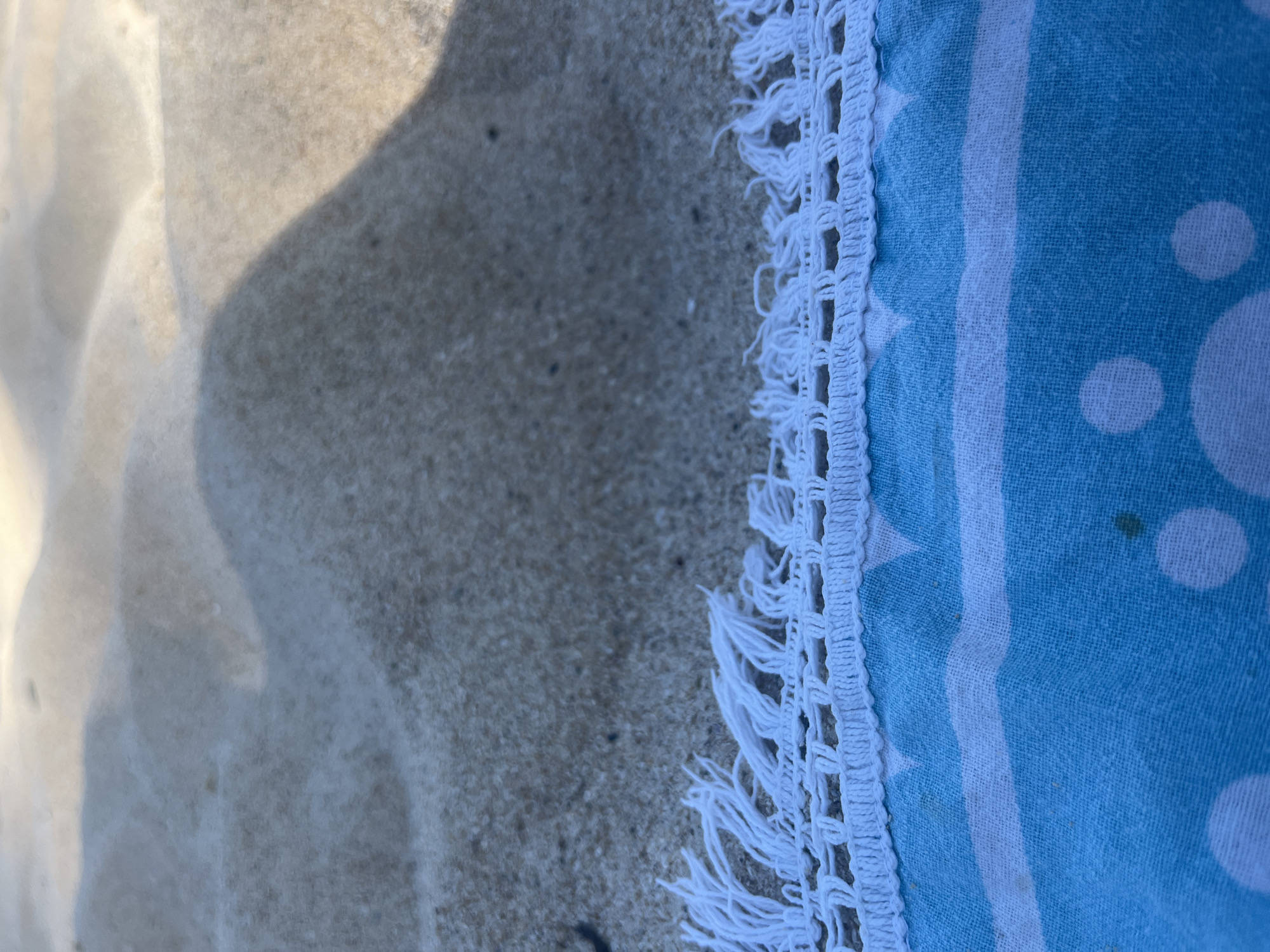 The image shows a close-up view of a patterned beach towel laid on sand. The towel is blue and white with a striped pattern and white polka dots. The top edge of the towel features white fringe and a crocheted border. The sand appears to be light brown and forms gentle dunes, with shadows cast across its surface, suggesting an outdoor environment, likely a beach. The image is taken from a low angle, focusing on the textures of the sand and the towel. There are no people or any other objects visible in the frame. The location is stated as Unknown, Unknown.