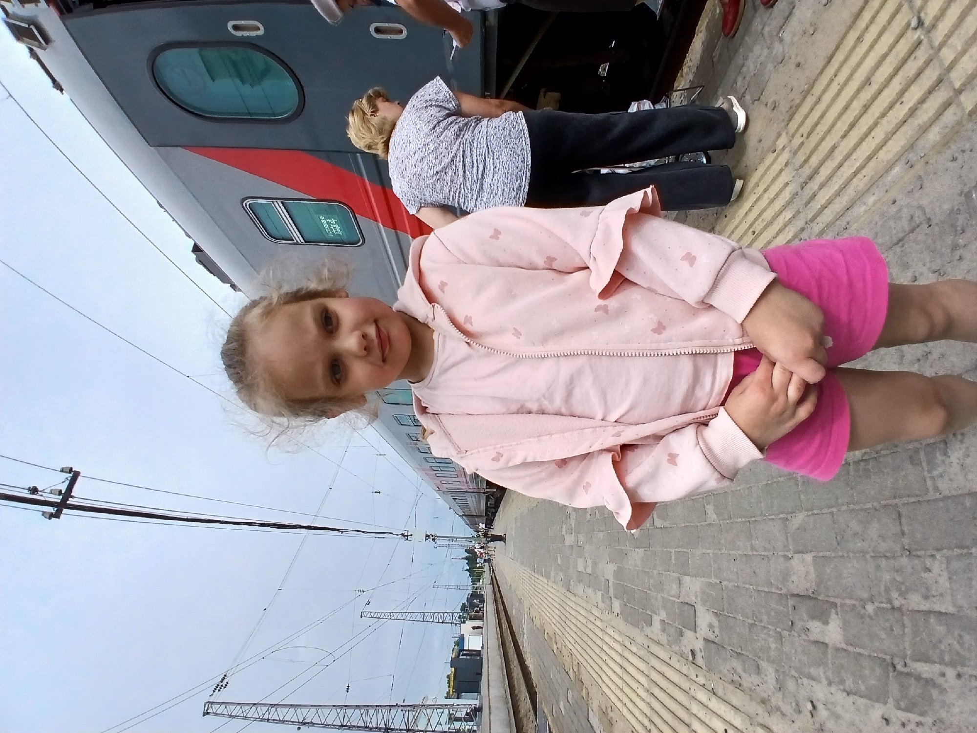 A young girl with blonde hair tied up, wearing a pink zip-up hoodie with butterfly patterns and a pink skirt, stands on a train platform. She is holding her hands clasped in front of her. Behind her, a grey train with a red stripe is parked. Two adult women are visible near the train; one is facing away from the camera, wearing a grey patterned top and dark pants, while the other is partially visible on the right, wearing dark pants and red shoes. The platform is paved with grey bricks and has yellow tactile paving. Overhead, power lines and utility poles crisscross the cloudy sky. In the background, more people and structures suggest a functioning train station, likely in Rtishchevo, Russia.