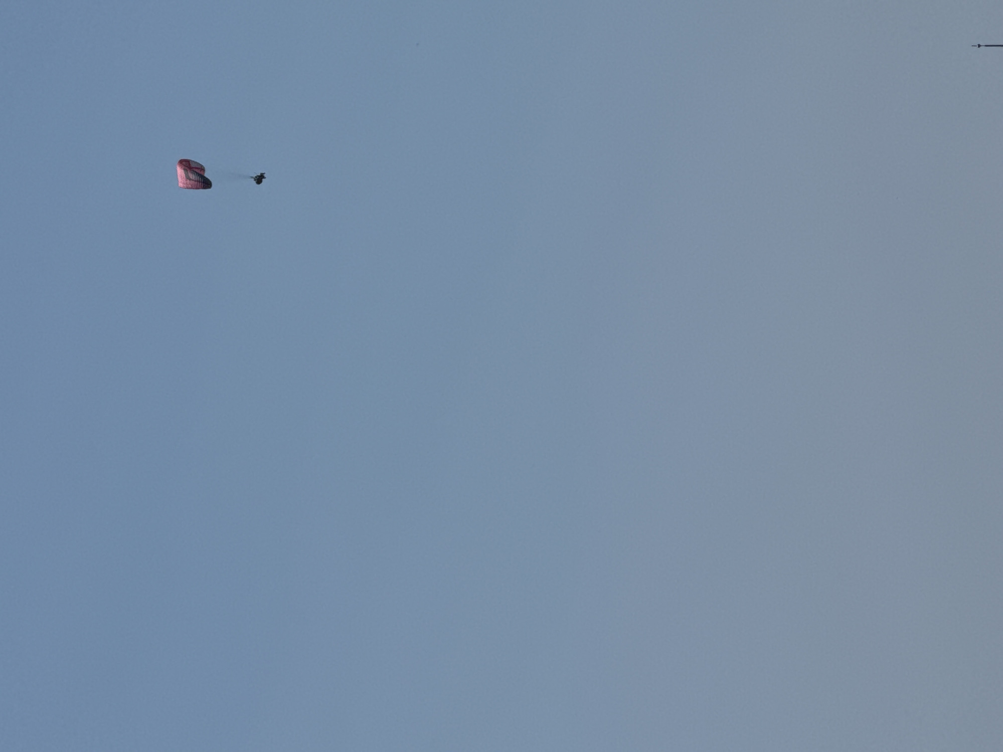 A parachute is descending in a clear blue sky. A person is attached to the parachute. The parachute appears to be red and black. A small trail of smoke or dust is visible behind the person. In the upper right corner of the image, there is a thin black line with a small dot at its end, possibly indicating a distant object or marker. The scene suggests an activity such as skydiving or a controlled descent. The lighting and sky color indicate it is daytime, with no clouds visible, suggesting fair weather. The location is given as Boreti, Montenegro. There is no visible text.