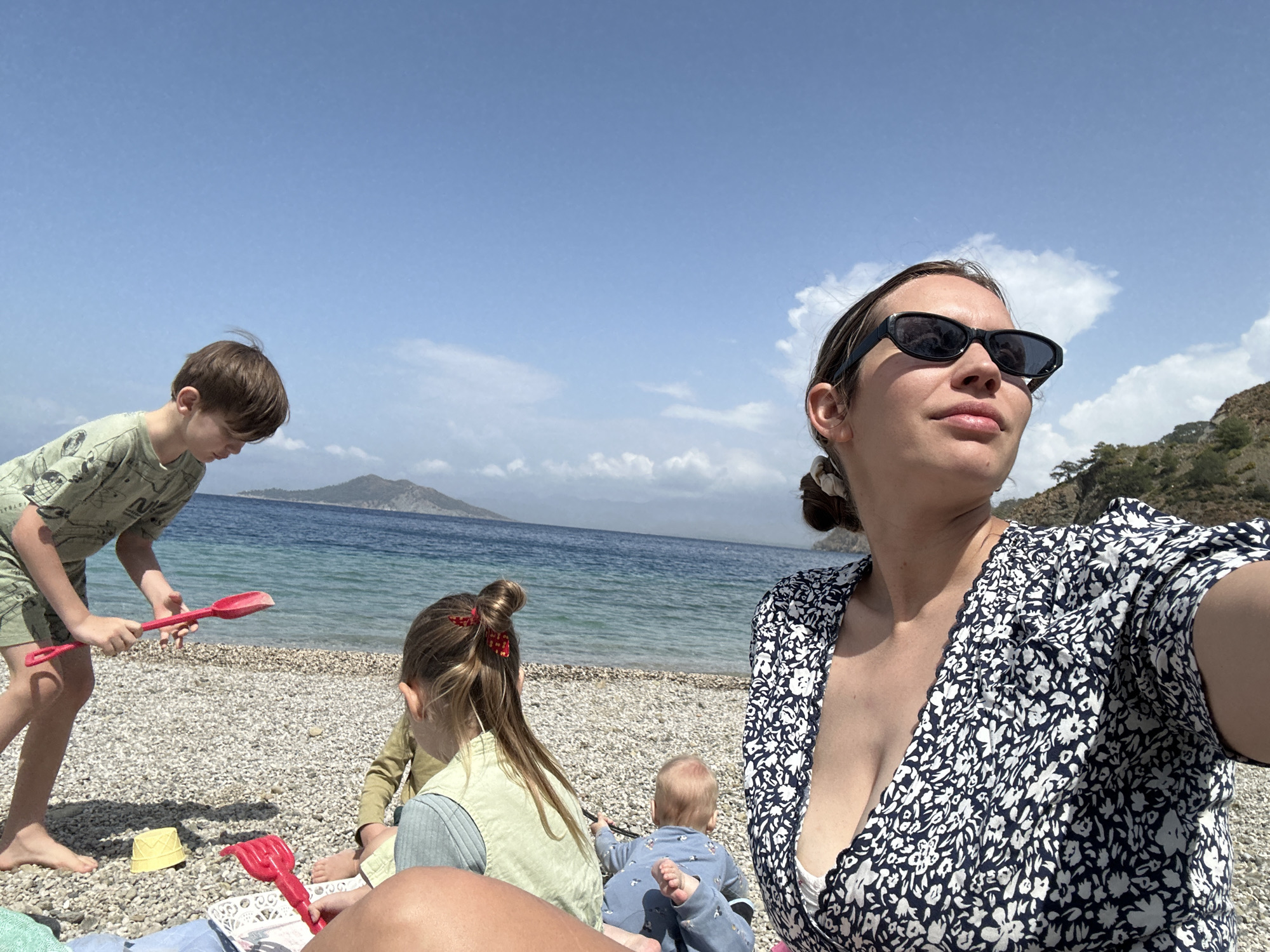 Captured on a beach in Fethiye, Turkey, the image presents a woman taking a selfie with three children nearby. The foreground includes the woman, her face angled to the right, clad in sunglasses and a floral-patterned dress. To her left, a boy in green shorts and shirt plays with a red toy shovel on the pebble-covered beach. Behind him, a girl with long hair tied with a red bow sits, and a baby in blue clothing lies on the ground. In the background, the Aegean Sea meets the sky, with a small island visible on the horizon. The sky is partly cloudy.