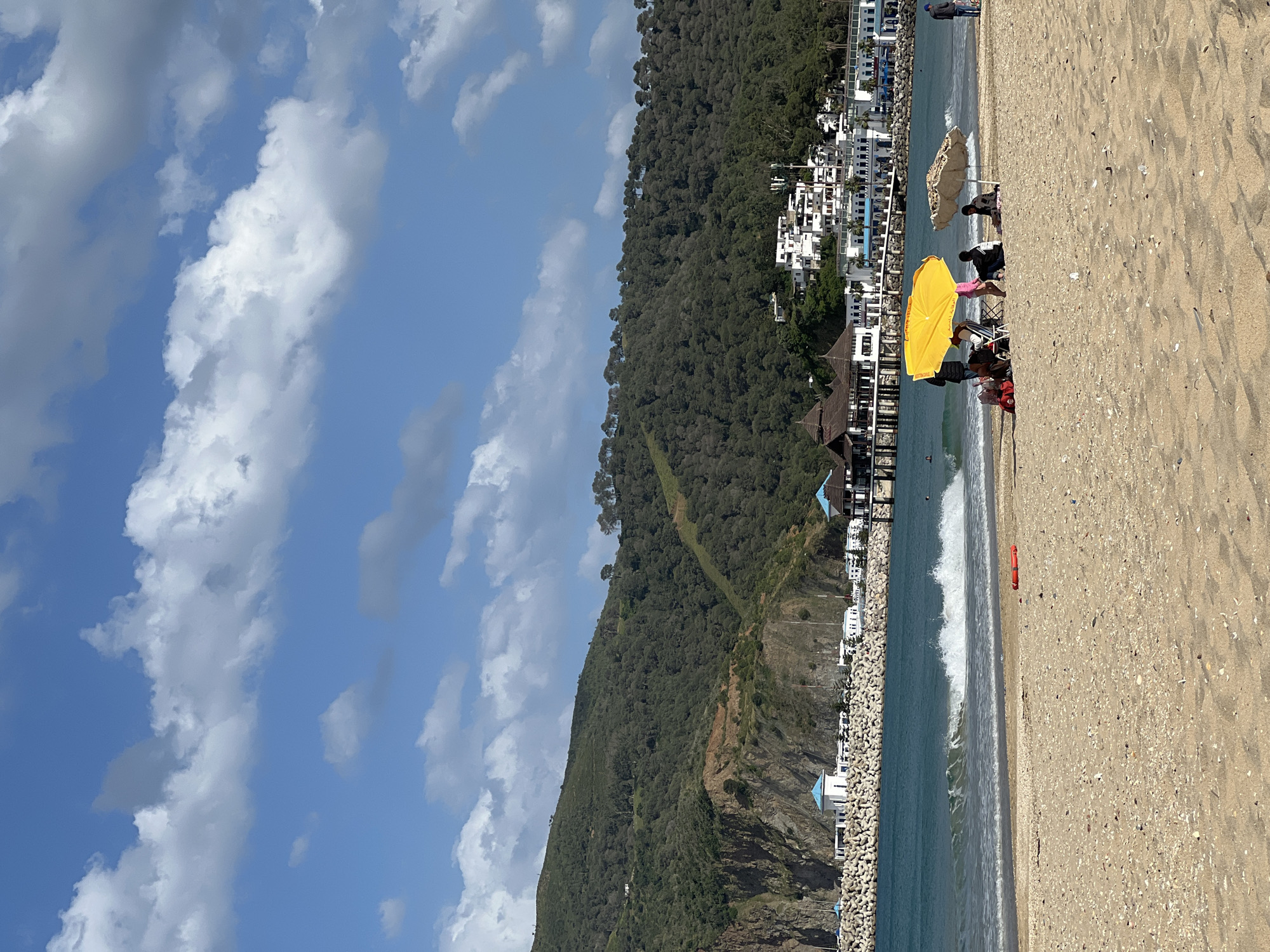 The image depicts a beach scene in M'Diq, Morocco, under a partially cloudy sky. A coastline with buildings lining the shore stretches along the center of the frame, backed by a tree-covered hillside. In the foreground, the beach is sandy, with a group of people gathered under a yellow umbrella. Other individuals are seated nearby under a second umbrella, and additional figures are visible further down the beach. A breakwater or series of concrete structures runs along the water's edge. The sky is blue with fluffy white clouds.