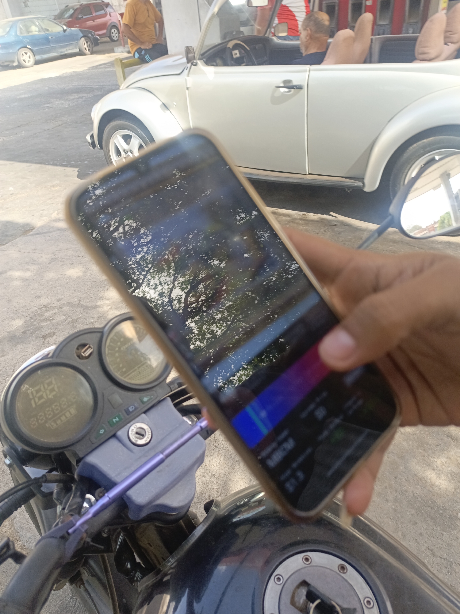 A person is holding a smartphone in front of a motorcycle dashboard. The smartphone screen shows a reflection of trees and a portion of a mobile application. The motorcycle is black, with a visible fuel tank cap and dashboard instruments showing "188888.8". In the background, partially out of focus, is a white convertible Volkswagen Beetle, parked on a paved surface. A man in an orange shirt is near the Beetle. Other vehicles are visible further in the background. The location is stated as La Tagua, Venezuela.