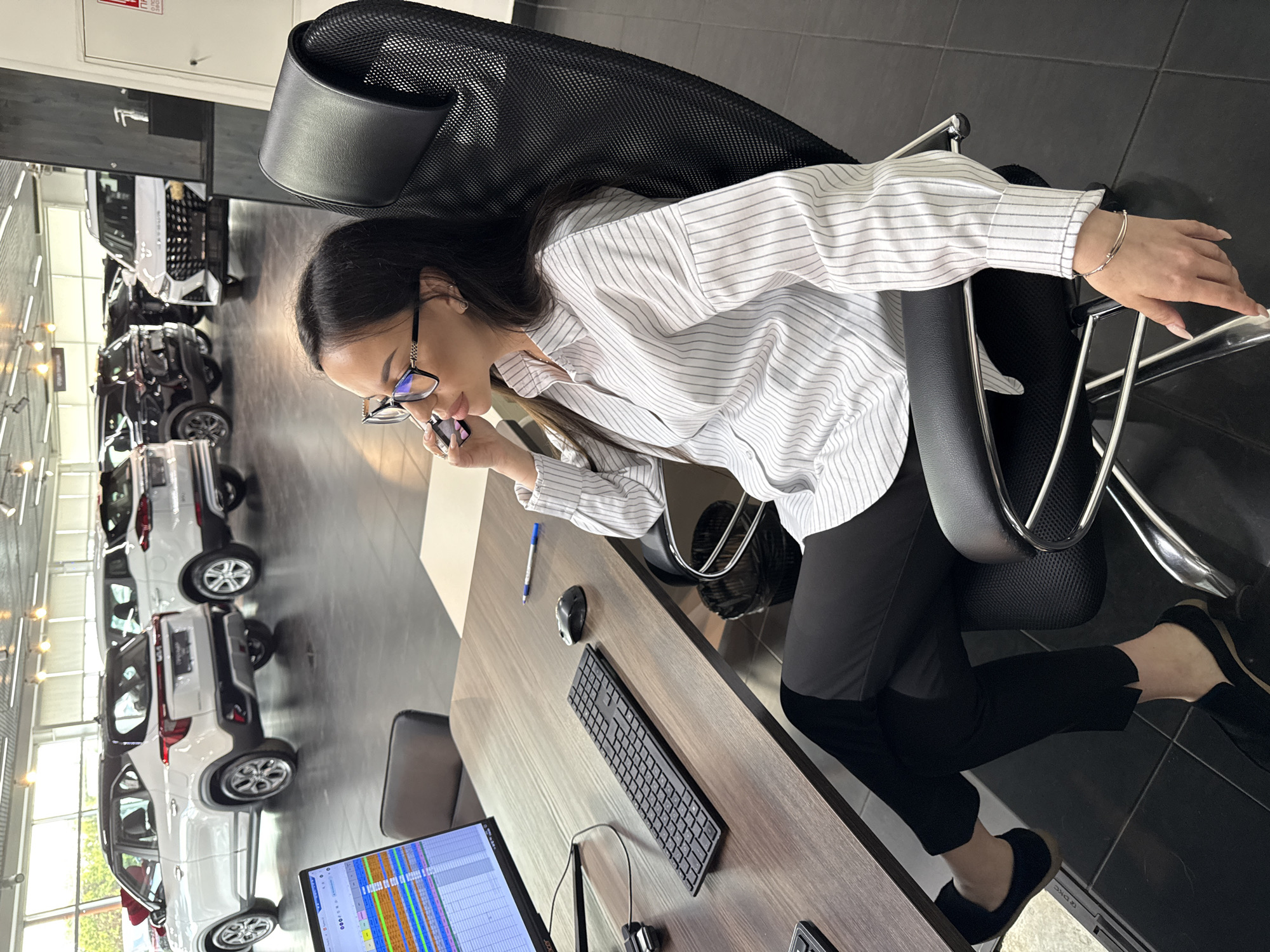 A woman wearing glasses, a pinstripe shirt, and black pants is seated in a black office chair in what appears to be a car dealership in Nizhny Novgorod, Russia. She is holding a mobile phone to her ear and talking. A dark brown wooden desk is in front of her, with a computer monitor displaying a spreadsheet, a keyboard, a computer mouse, and a pen. Several cars, including white SUVs and a black SUV, are visible in the background through large windows. The floor is tiled in black and white.