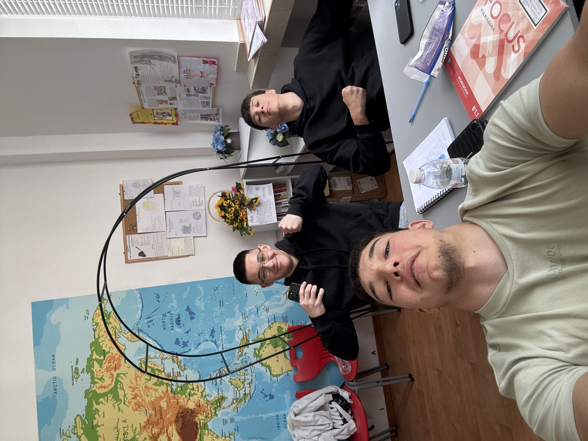 Three young men are captured in a selfie taken in what appears to be a classroom or study environment in Zlatograd, Bulgaria. The man closest to the camera, positioned on the right side of the frame, has light facial hair and is wearing a light green t-shirt with the text "ALTITUDE" visible. He is looking directly into the camera with a slight smirk.
Behind him, two other young men, both wearing black hoodies, are seated at desks. The one closer to the center is smiling and holding a black smartphone. The other, further back, is also looking towards the camera. The overall mood is casual and friendly.
The setting includes a large physical world map displayed on the wall to the left, prominently featuring landmasses and oceans such as "ARCTIC OCEAN", "NORTH PACIFIC OCEAN", "MONGOLIA", "AUSTRALIA", and "INDIAN OCEAN". On the right side, a desk holds various items: a spiral notebook with handwritten notes, a plastic water bottle, a blue pen, a black phone, and a textbook titled "FOCUS FOR BULGARIA" with "WORKBOOK" and "B1.1" also visible. An "OREO" wrapper is also on the desk.
Above the desks, a bulletin board displays papers, including one with "Mein Traumjob" written on it. Shelves with books and binders, as well as decorative floral arrangements (blue and yellow flowers), are also visible in the background. The room has white walls and a wooden floor, with red chairs partly visible.