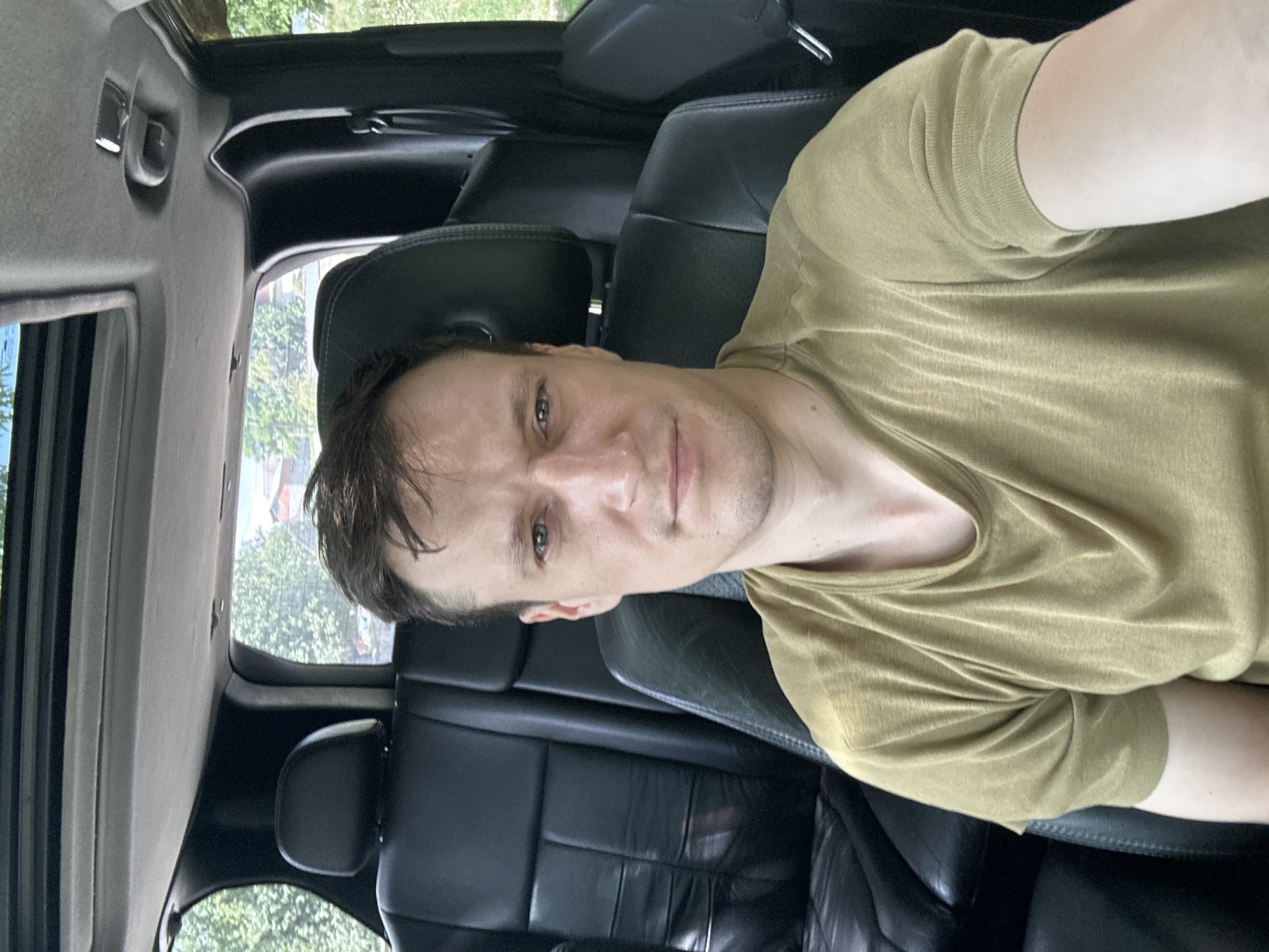 In Makiv, Ukraine, a man is photographed from the chest up, seated in the driver's seat of a car. The car's interior is visible, with black leather seats and a sunroof. Outside, trees and houses can be seen through the car windows. The man has fair skin, short brown hair, and is wearing a plain olive-colored t-shirt. His gaze is directed towards the camera. The lighting suggests it is daytime.
